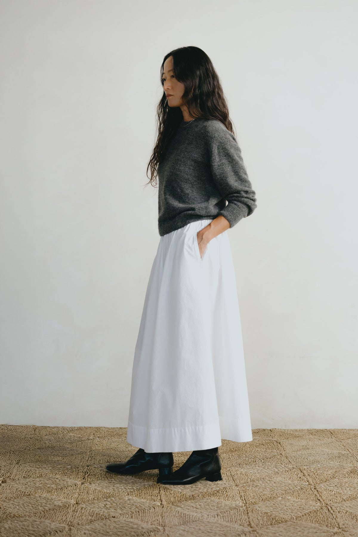 A woman stands in profile wearing a dark gray knit sweater, a long white skirt with side pockets, and black ankle boots, set against a plain off-white wall and a woven straw floor.