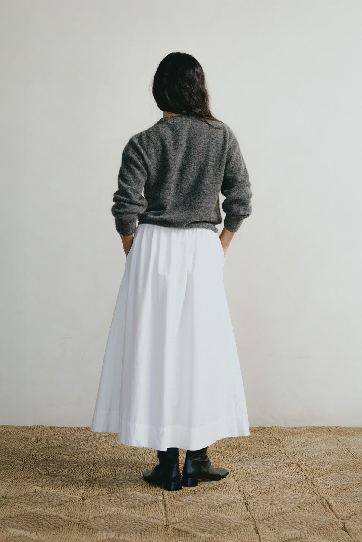 A woman is shown from behind wearing a dark gray knit sweater and a long white skirt, standing on a textured woven rug against a plain off-white wall.