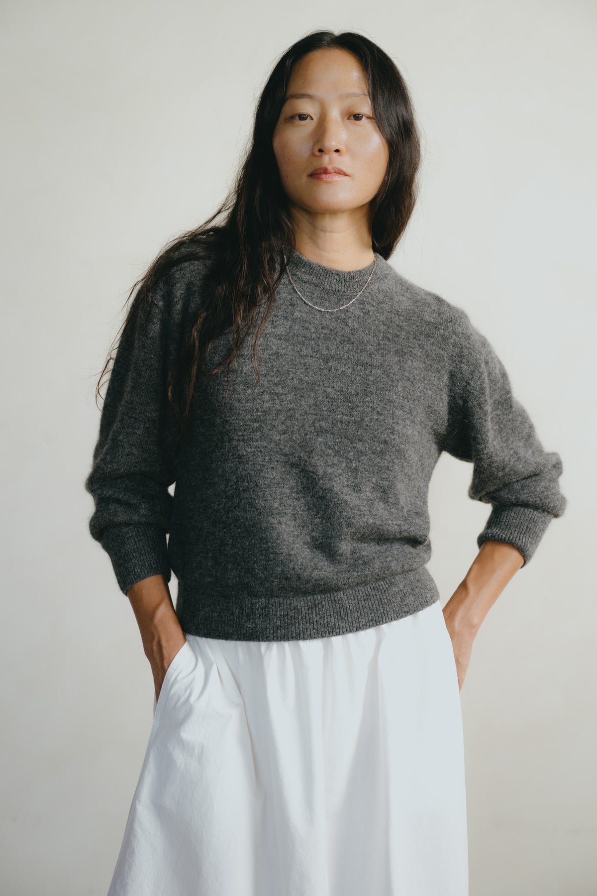 A woman wearing a dark gray knit crewneck sweater and a white skirt, standing against a plain light gray background.