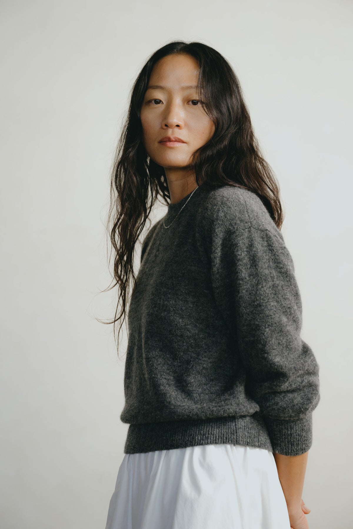 A woman with long dark hair wearing a dark gray knit sweater and a white skirt, photographed against a plain light gray background.