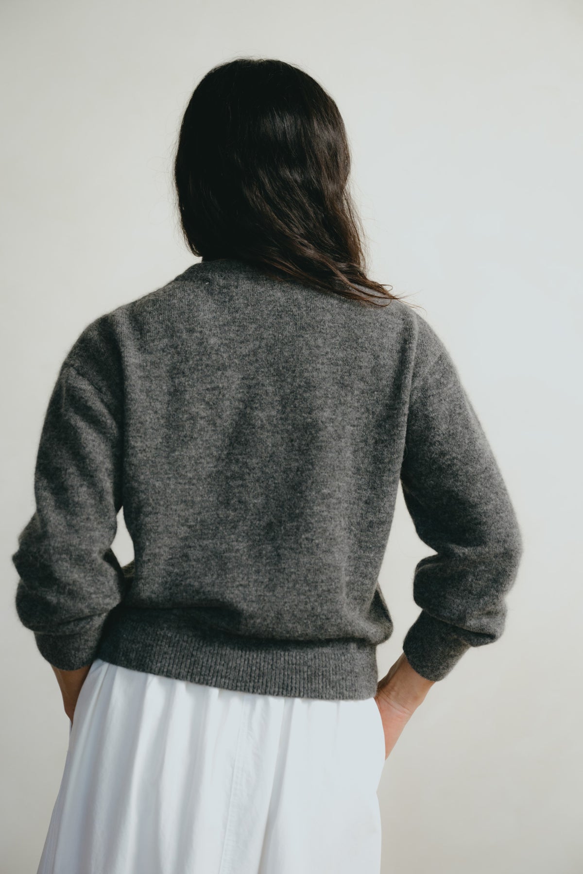 A woman viewed from behind wearing a dark gray knit sweater and a white skirt, standing against a plain light gray wall.
