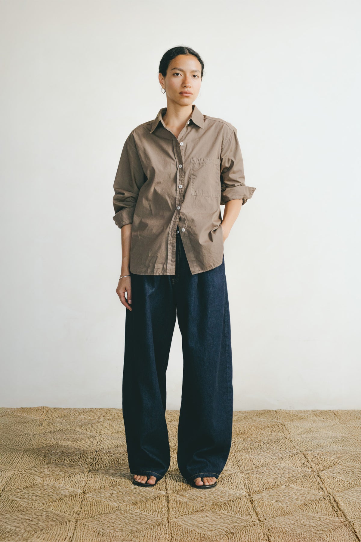 A model stands in a minimalist studio setting wearing a brown button-down shirt with rolled-up sleeves and dark wide-leg trousers.