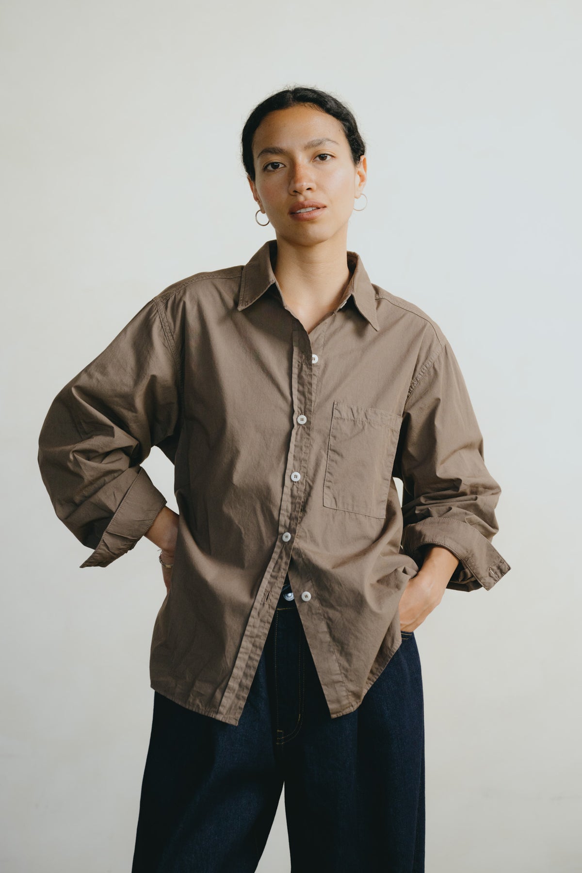 A model wearing an oversized, long-sleeve brown button-up shirt with a chest pocket, paired with dark blue jeans, standing against a plain light gray background.
