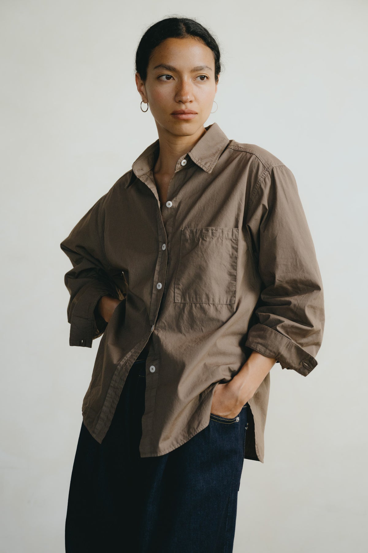 A woman wearing an oversized brown button-up shirt with rolled sleeves and dark blue jeans, standing against a plain light gray background.