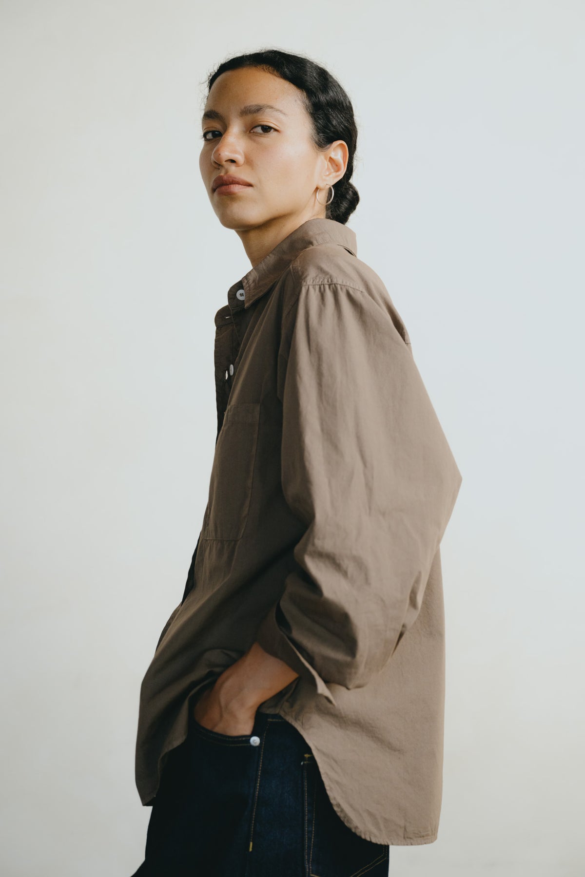 A woman wearing an oversized taupe-brown button-up shirt and dark blue jeans, standing against a plain light gray background.