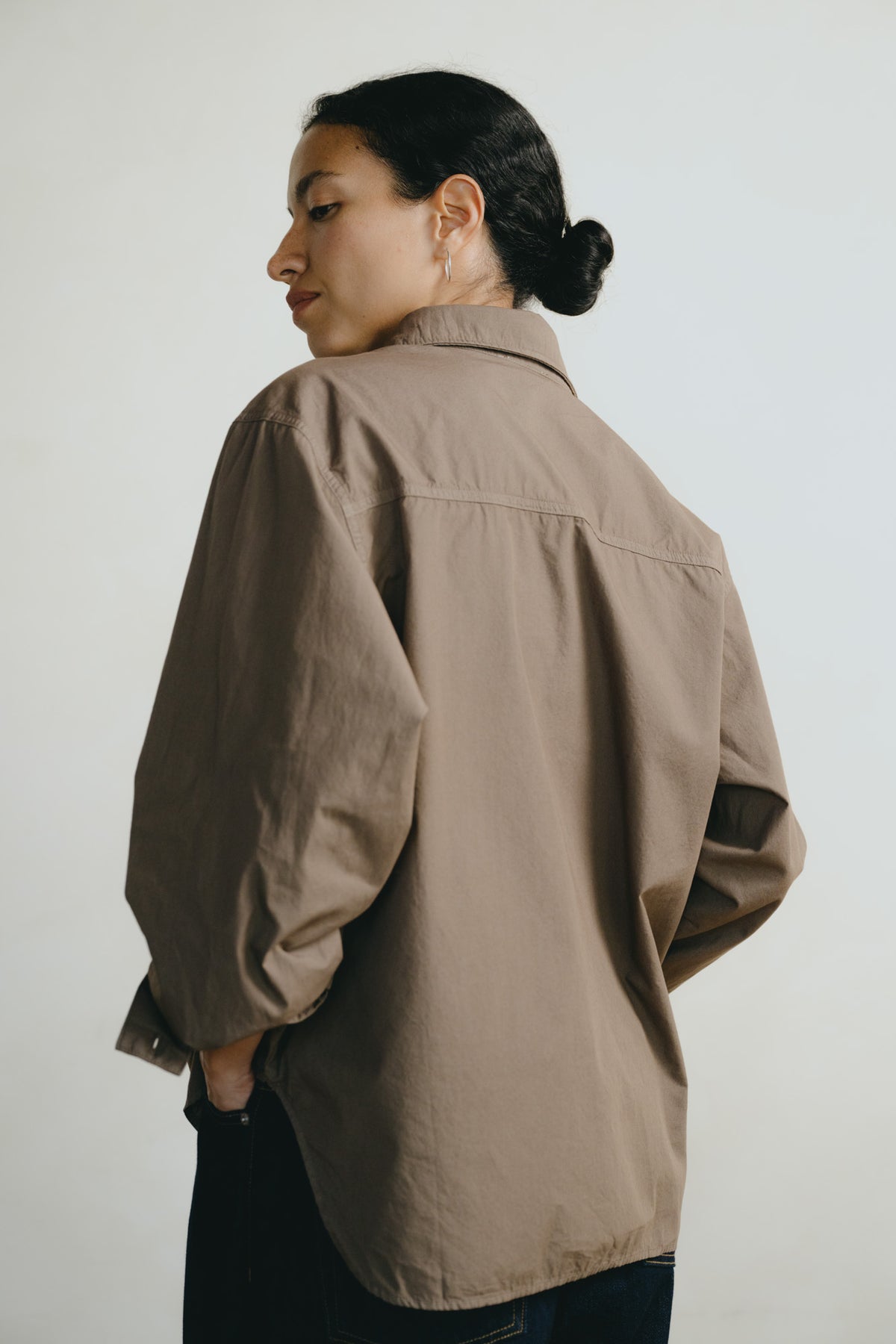 A woman shown from the back wearing an oversized taupe-brown utility shirt over a dark top, photographed against a plain light gray studio background.