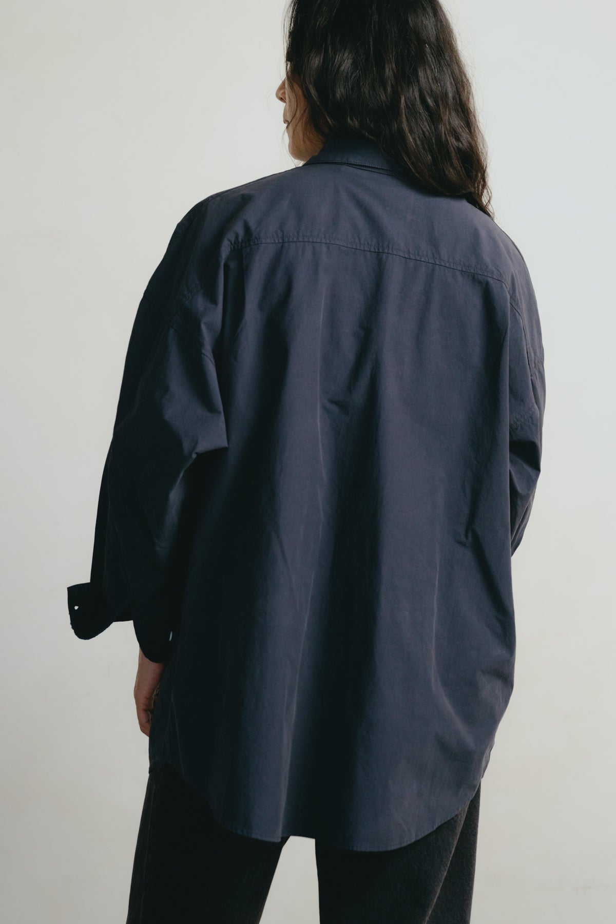 A person shown from the back wearing an oversized dark navy button-up shirt and black pants against a plain light gray studio background.