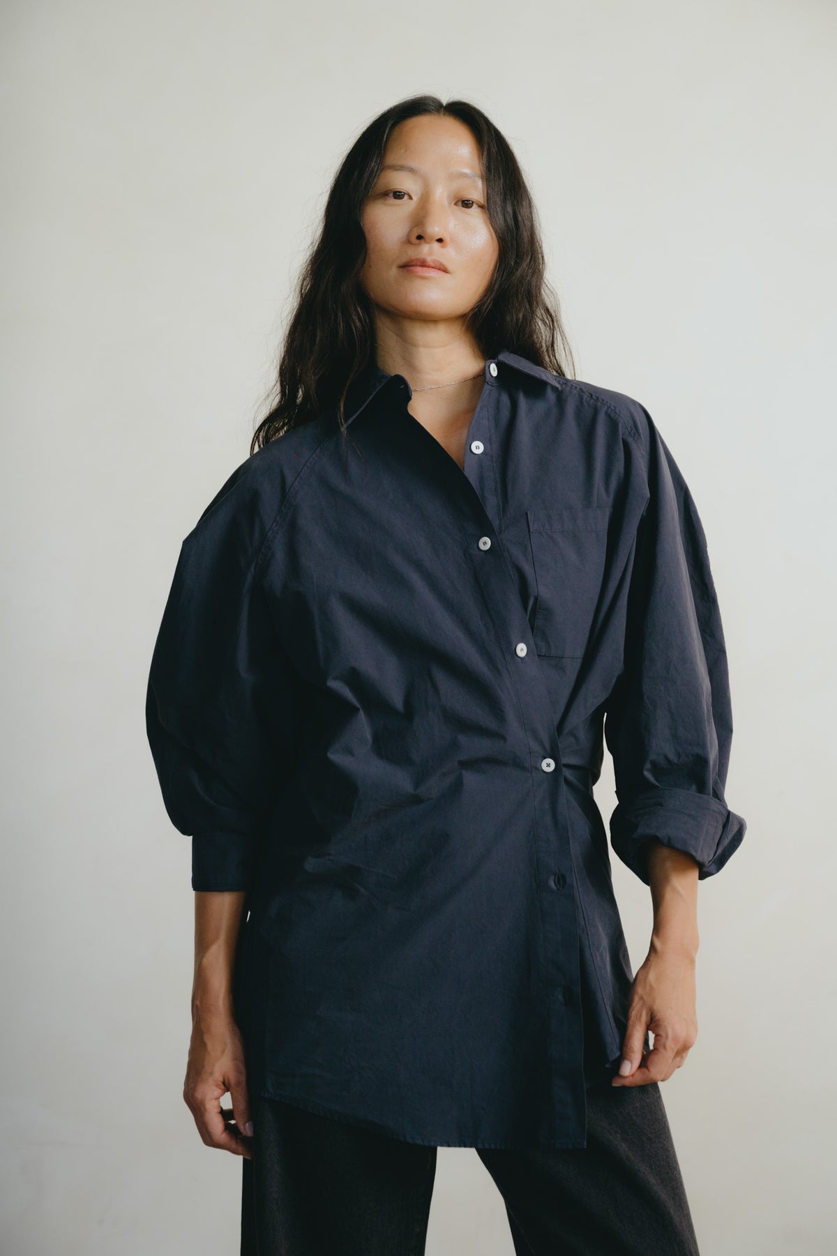A woman modeling a dark navy blue button-down shirt dress with a tie waist and voluminous sleeves, set against a plain light gray background.