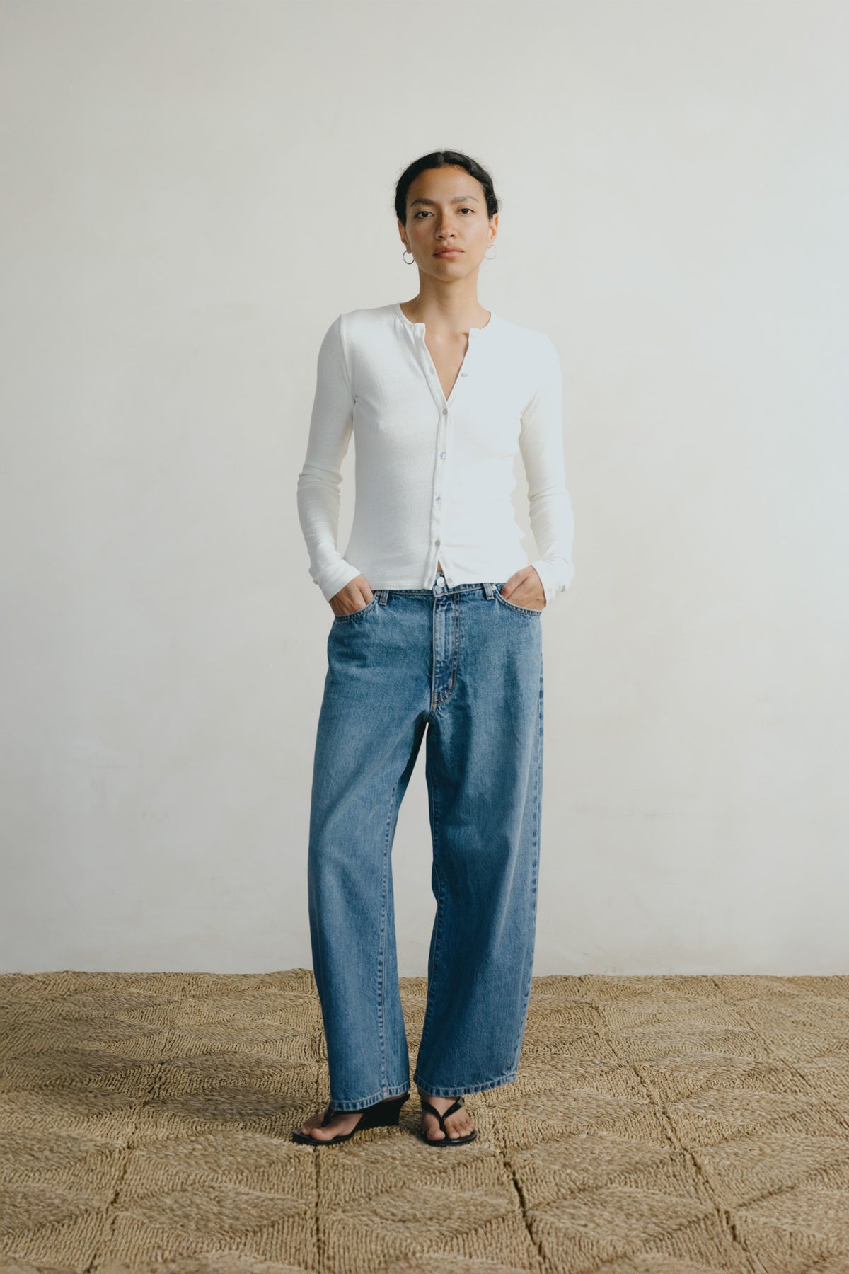A woman stands in a studio wearing a white long-sleeve button-front top, wide-leg blue jeans, and dark sandals against a plain light wall and woven floor.