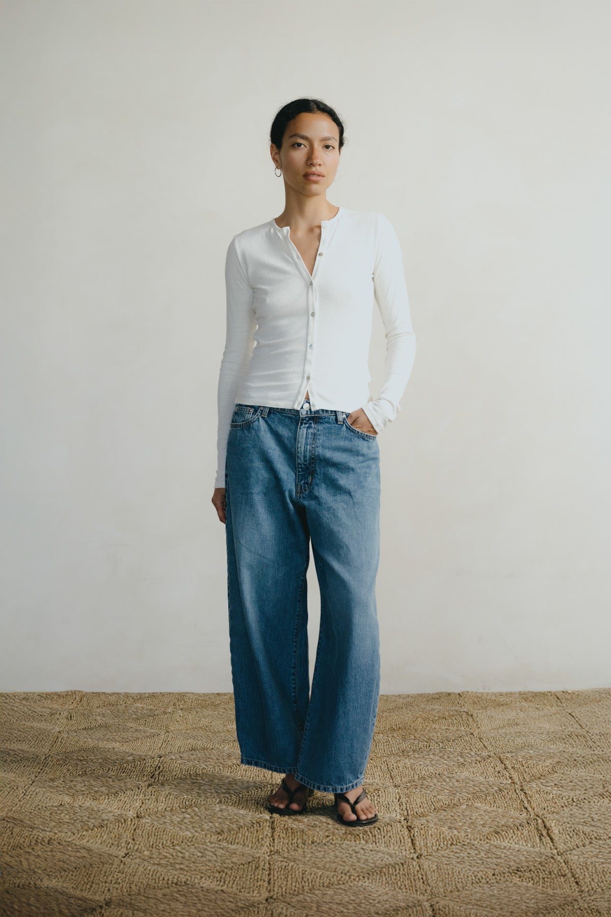A model stands against a plain off-white wall wearing a fitted white long-sleeve button-front top and wide-leg blue jeans, with dark sandals, on a woven straw floor.