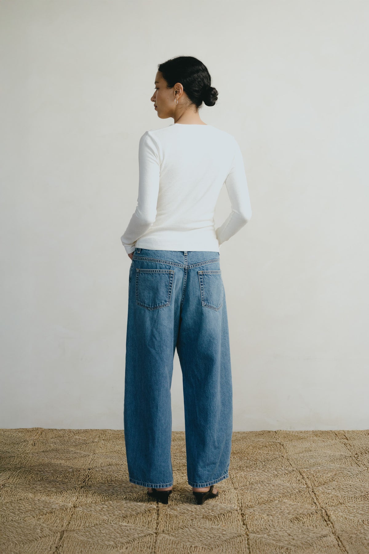 A woman stands with her back to the camera wearing a fitted white long-sleeve top and wide-leg blue denim jeans, standing on a woven straw rug against a plain off-white wall.