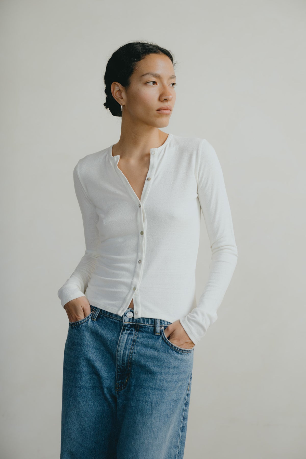 A woman wearing a fitted white long-sleeve button-front top and blue jeans, standing against a plain light gray studio background.