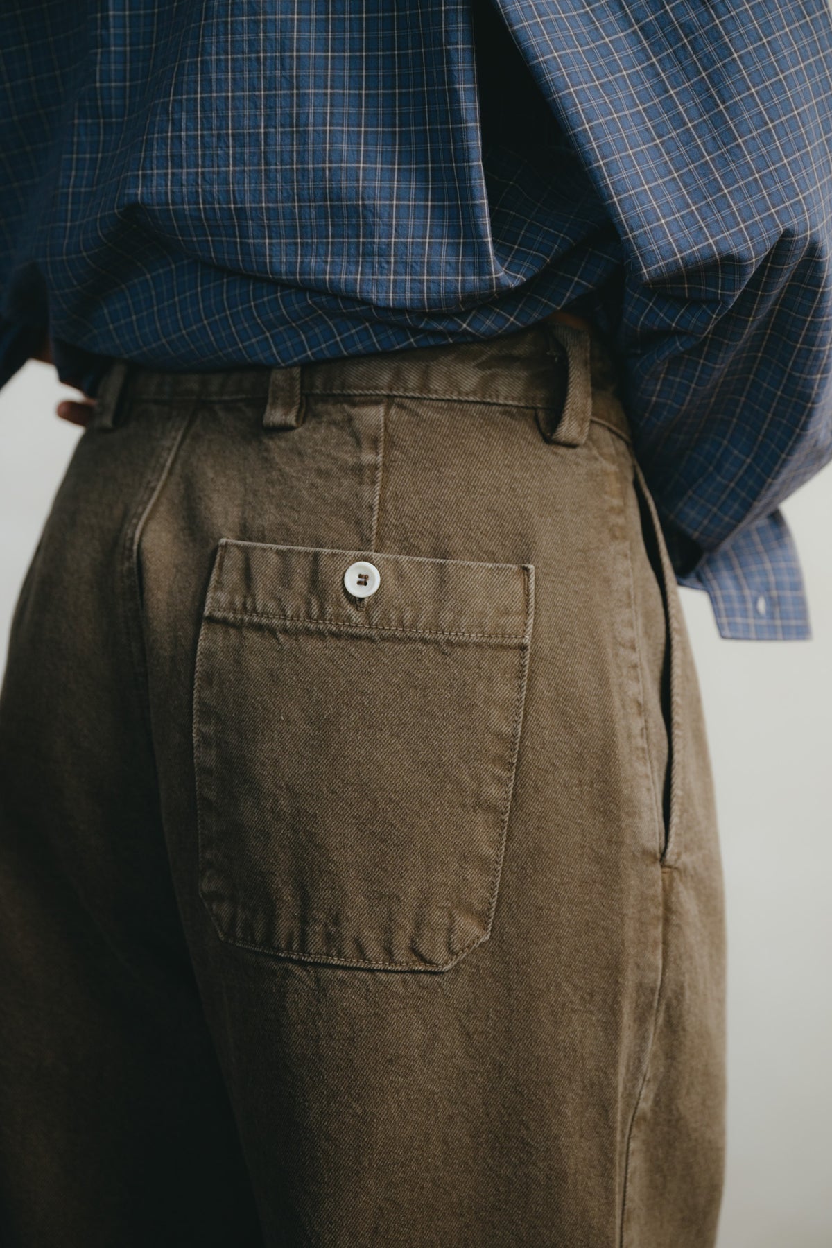 Close-up of a person wearing brown canvas trousers with a back pocket and a blue plaid shirt tucked in, photographed against a plain light background.