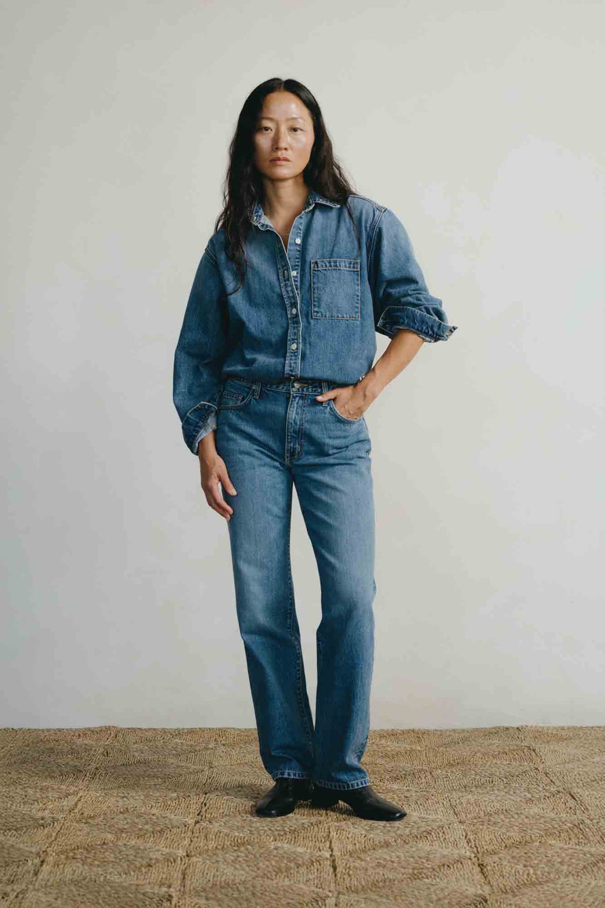 A model stands in a studio wearing a matching medium-wash blue denim shirt and jeans, paired with black shoes, against a light gray wall and a textured tan floor.