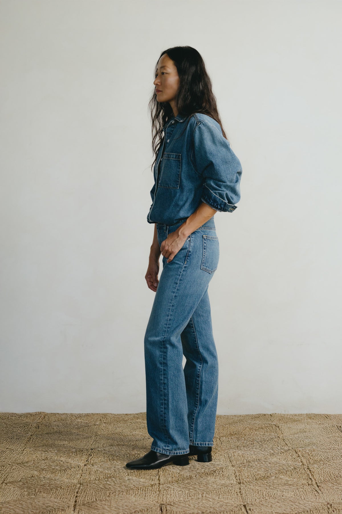 A woman stands in profile wearing a blue denim jacket with voluminous sleeves and matching blue denim jeans, paired with black shoes, against a plain off-white wall and a woven jute floor.