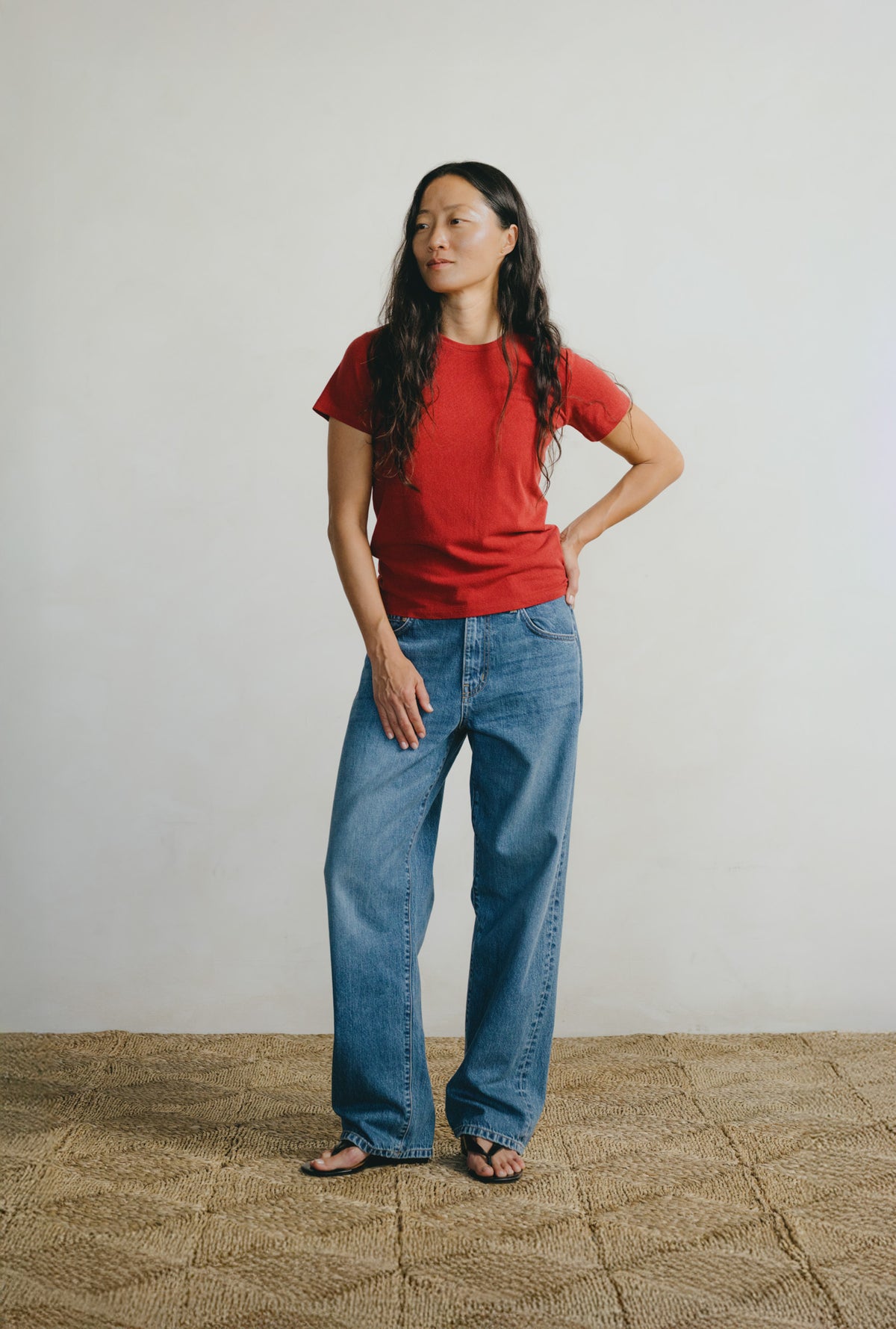 A woman stands in a studio wearing a red short-sleeve crewneck t-shirt and wide-leg blue jeans, paired with black sandals. She is posed against a neutral off-white wall and a textured beige woven floor.
