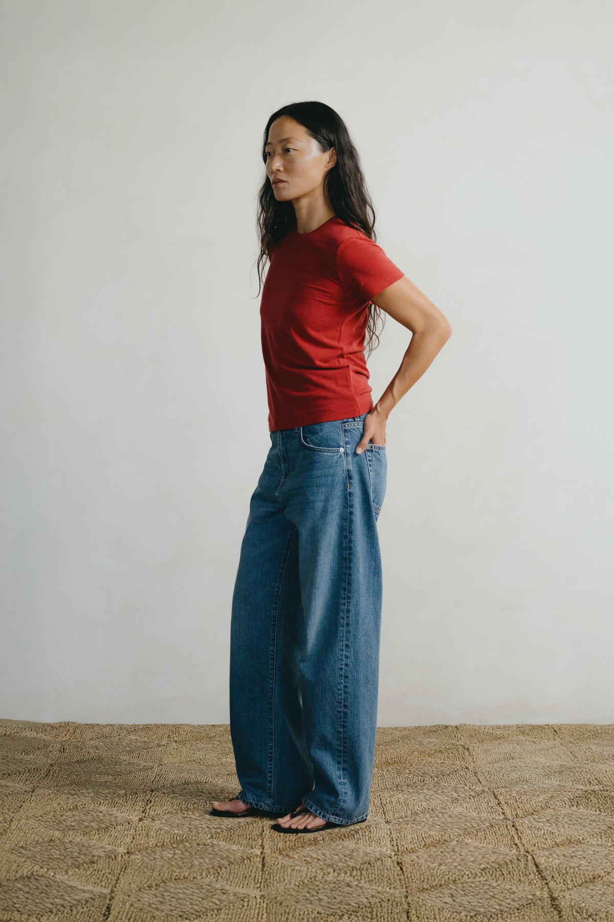 A woman stands in profile wearing a red short-sleeve crewneck t-shirt and wide-leg blue jeans, standing on a woven straw rug against a plain off-white wall.