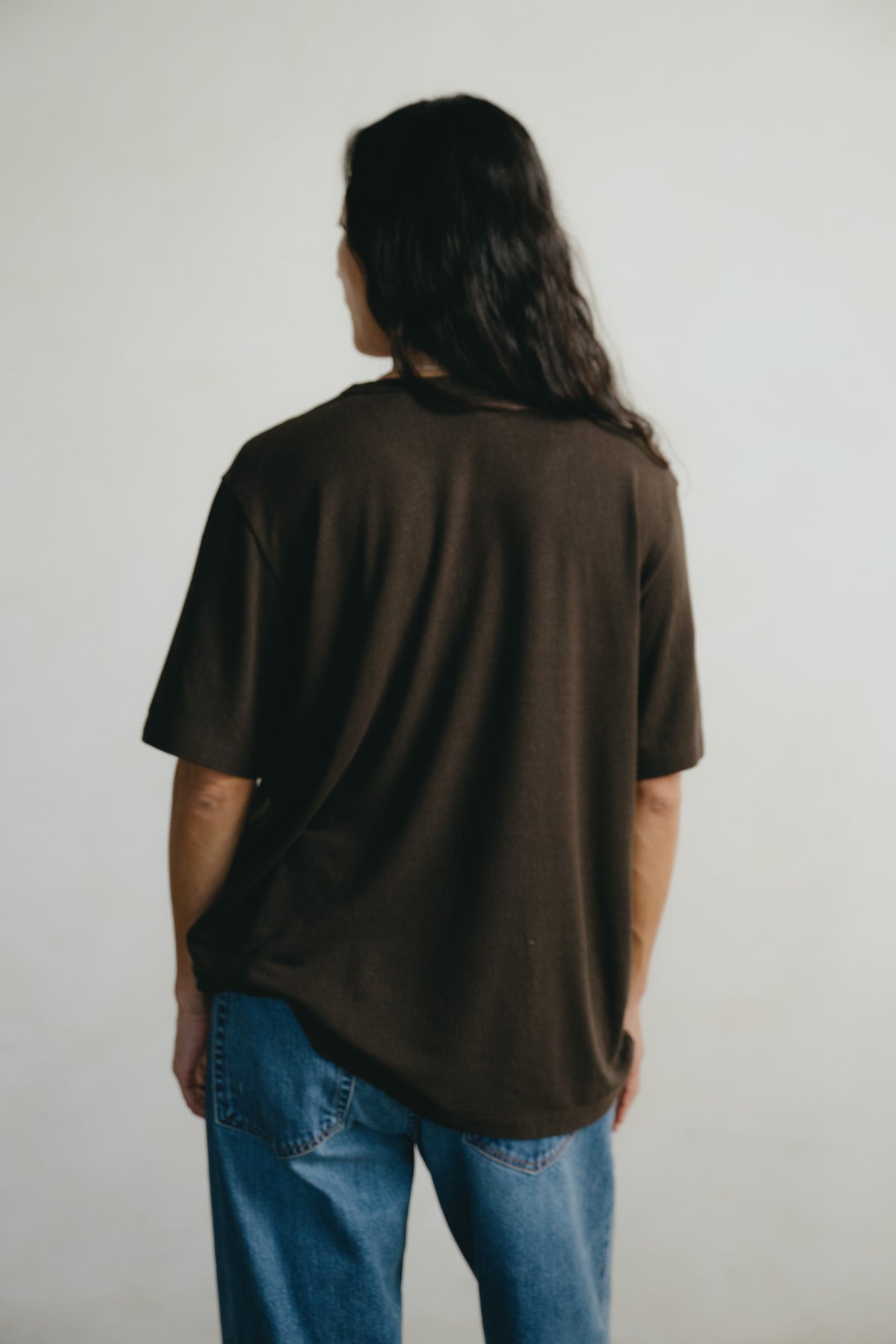 A person viewed from behind wearing an oversized dark brown short-sleeve t-shirt and blue jeans, standing against a plain light gray background.