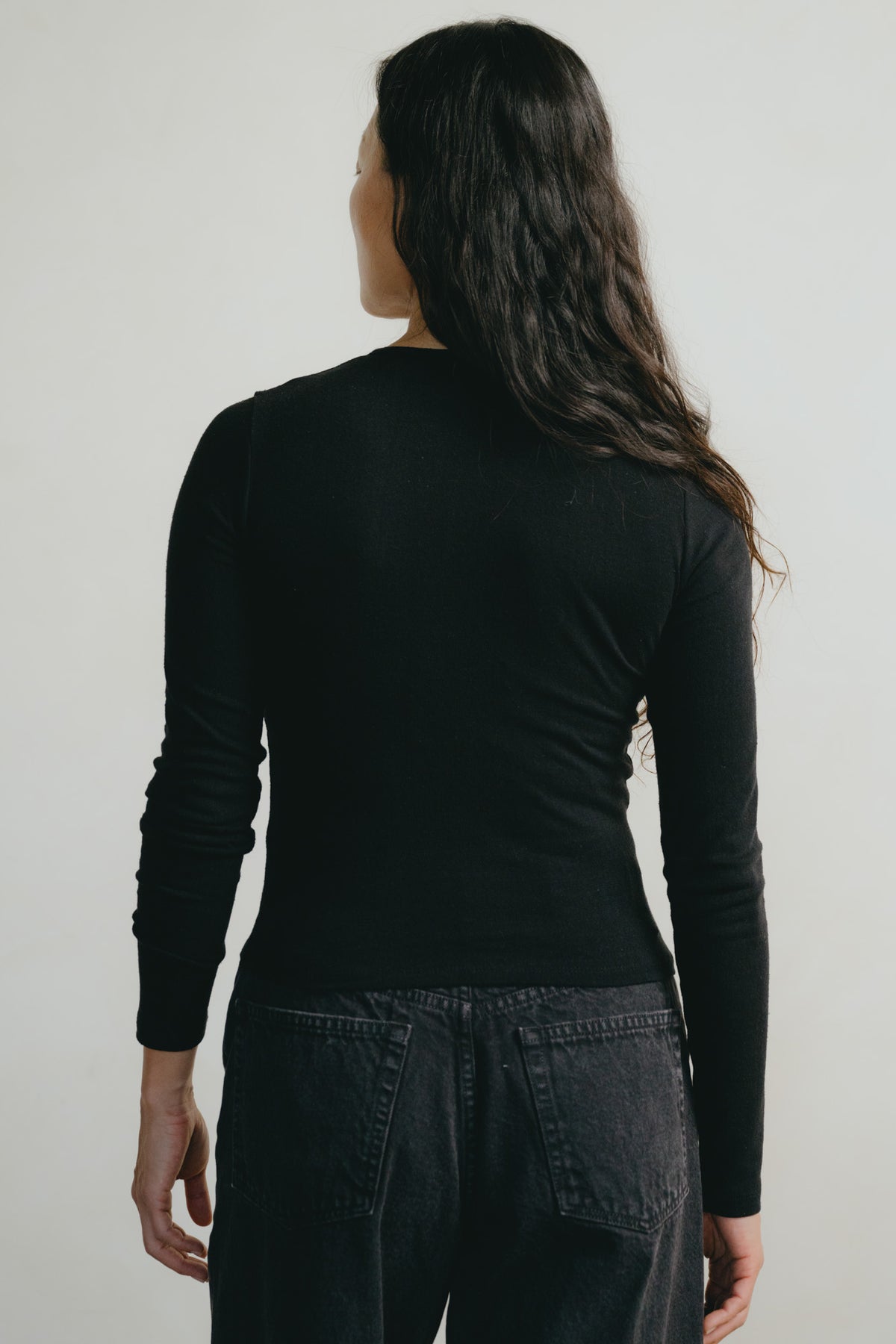 A woman with long dark hair is shown from behind wearing a fitted black long-sleeve top and dark jeans against a plain light gray studio background.