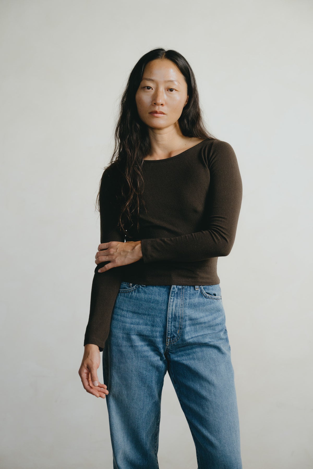 A woman with long dark hair stands against a plain light gray studio background, wearing a fitted dark brown long-sleeve top and blue jeans.