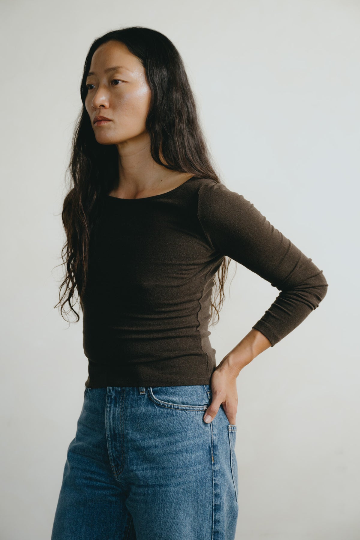 A woman wearing a dark brown long-sleeve boat-neck top and blue jeans, standing against a plain light gray background.