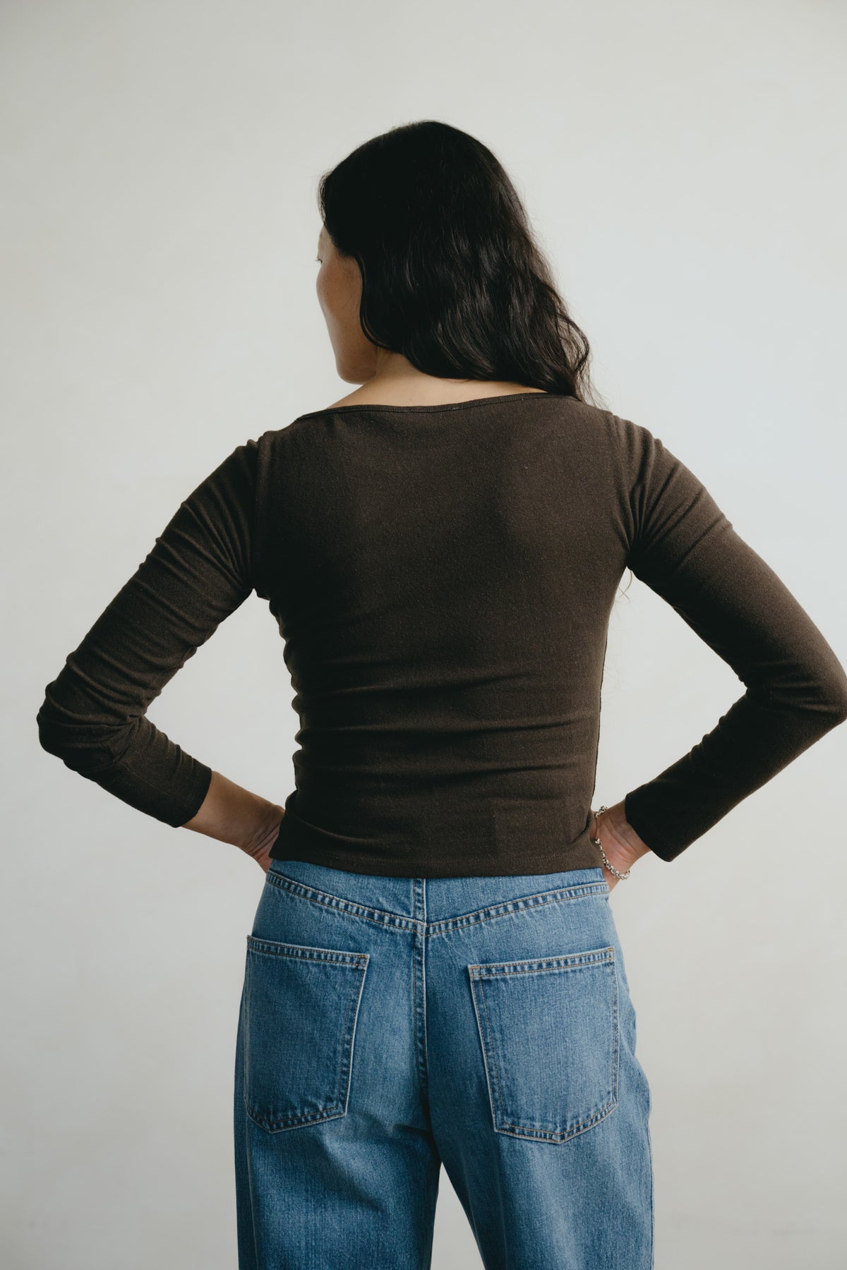 A woman shown from behind wearing a dark brown long-sleeve boat-neck top and blue jeans, standing against a plain light gray background.