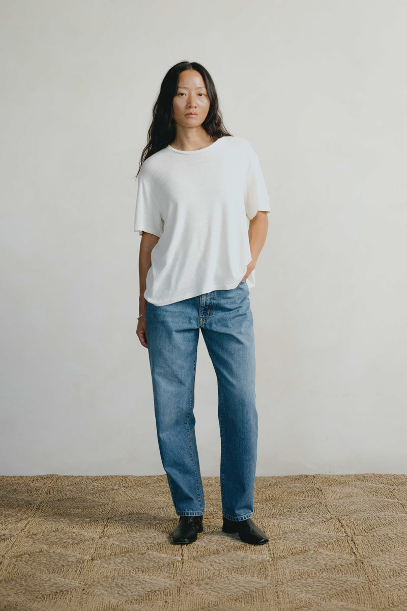 A woman stands in a studio wearing a loose white short-sleeve T-shirt, blue straight-leg jeans, and black shoes against a plain light wall and woven floor.