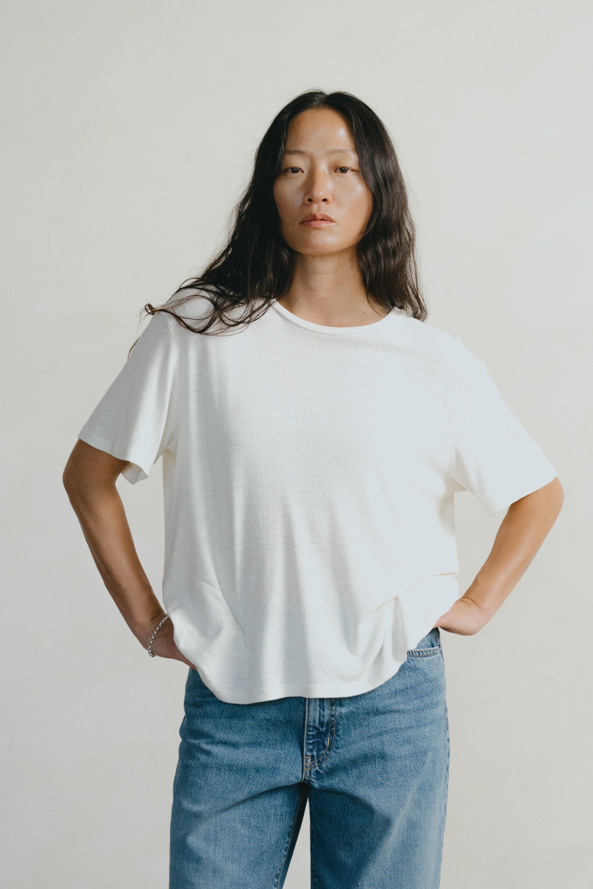 A woman modeling a loose-fitting white short-sleeve t-shirt with blue jeans, standing against a plain light gray studio background.