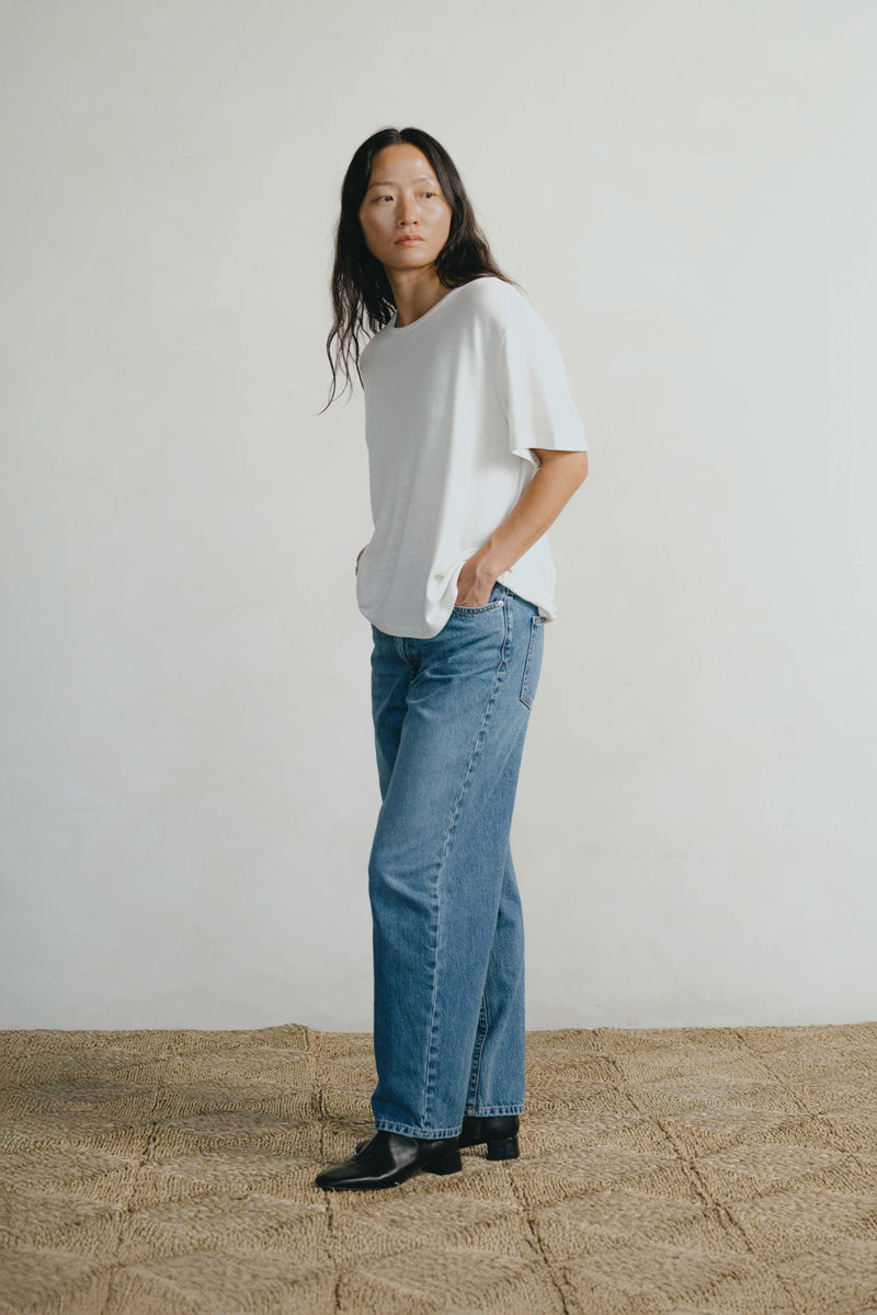 A woman stands in a studio wearing a loose white short-sleeve T-shirt, light blue wide-leg jeans, and black shoes against a plain off-white wall and woven floor.