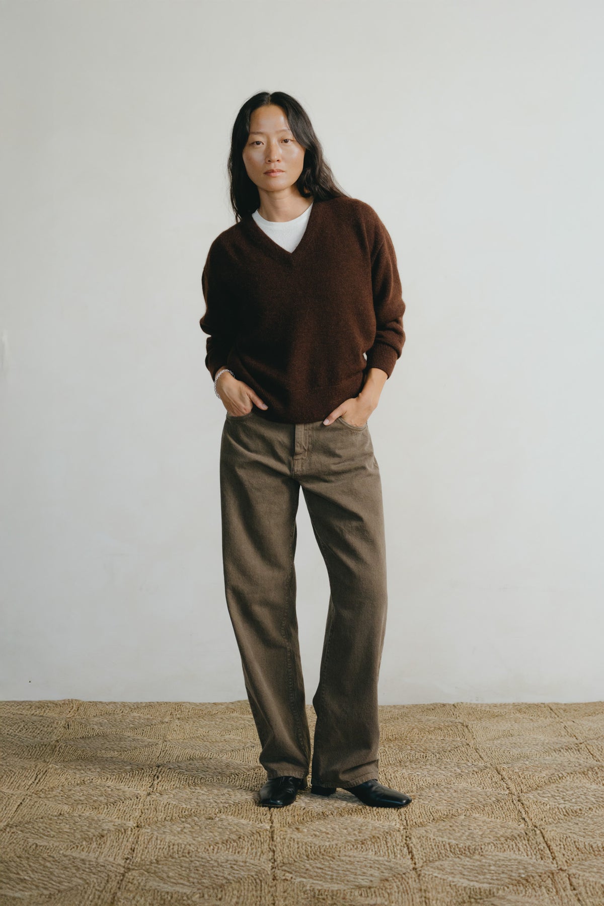 A model stands against a plain off-white wall wearing a dark brown v-neck knit sweater over a white t-shirt, paired with brown wide-leg trousers and black shoes.