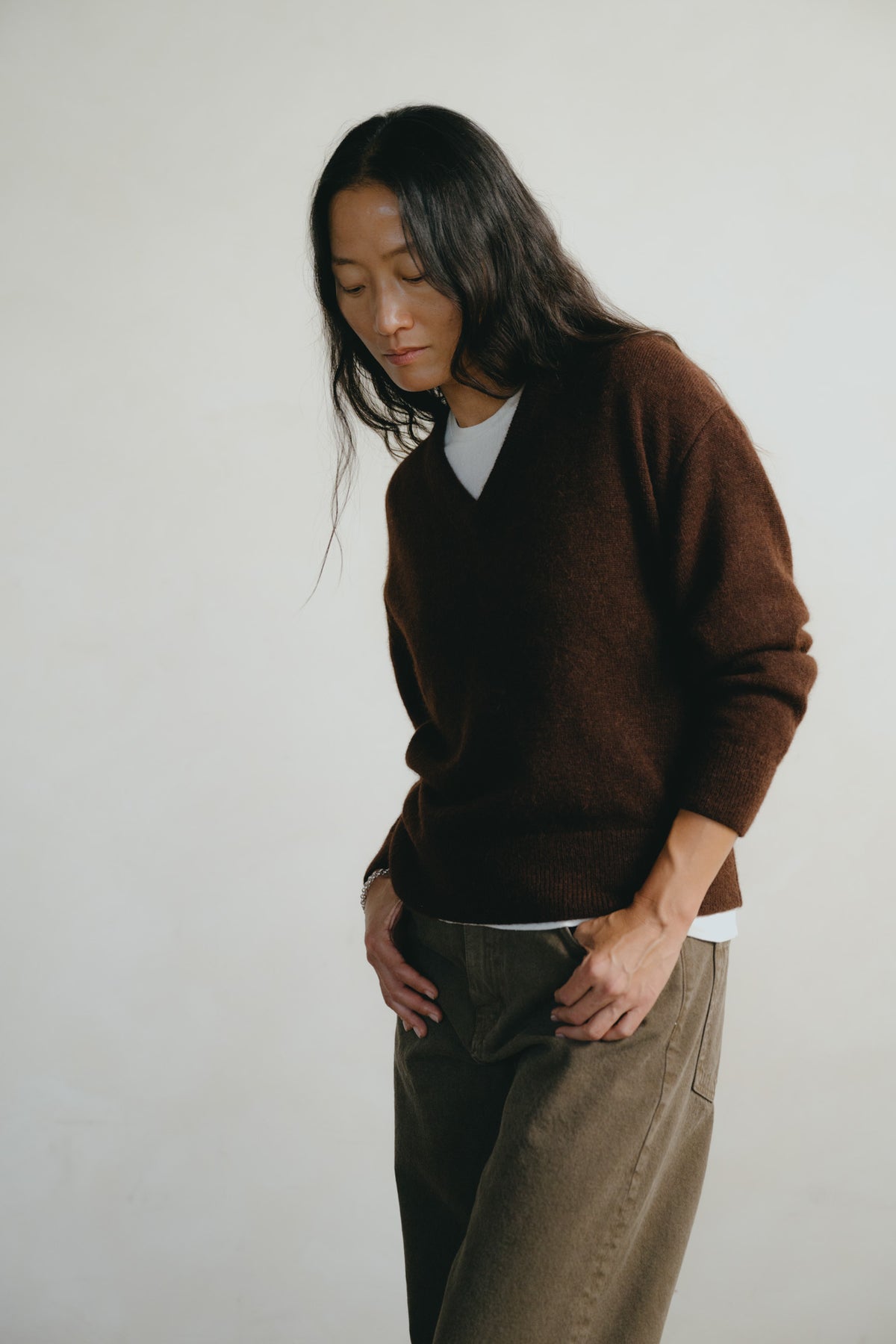 A woman with long dark hair is shown from the waist up, wearing a brown V-neck knit sweater over a white undershirt and brown trousers, standing against a plain off-white background.