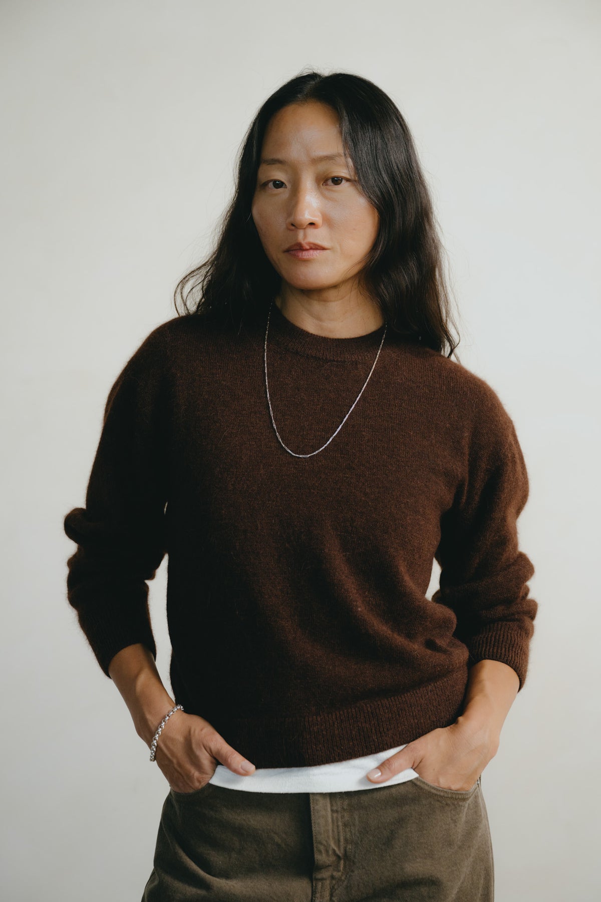 A woman with long dark hair wearing a dark brown knit crewneck sweater, a silver chain necklace, and olive green corduroy pants, standing against a plain light gray background.