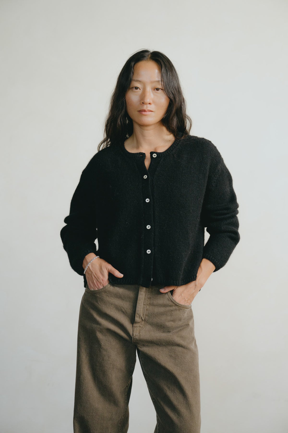 A woman wearing a black button-up knit cardigan and brown wide-leg pants, standing against a plain light gray studio background.
