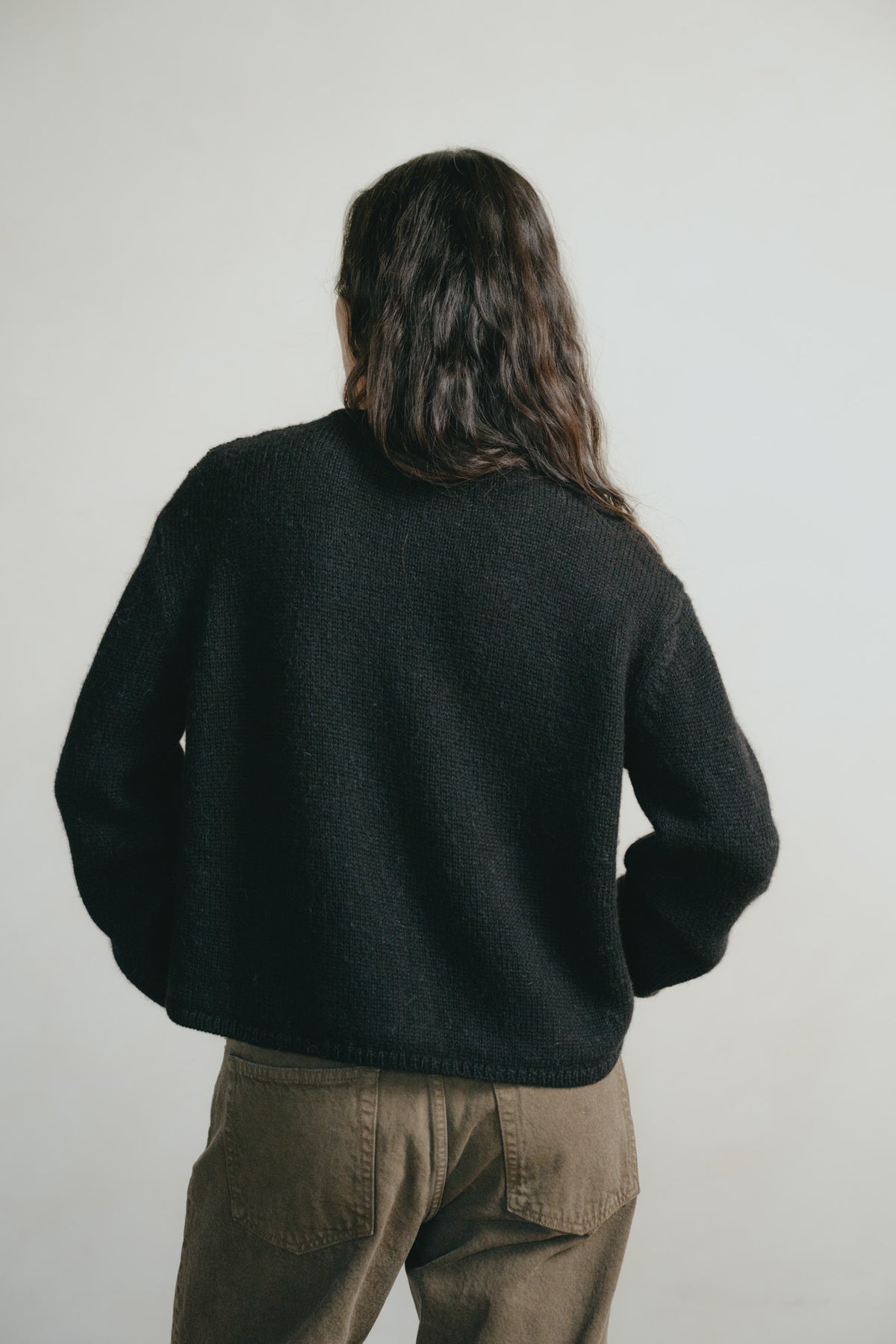 A person viewed from behind wearing a dark charcoal knit sweater and brown pants against a plain light gray background.