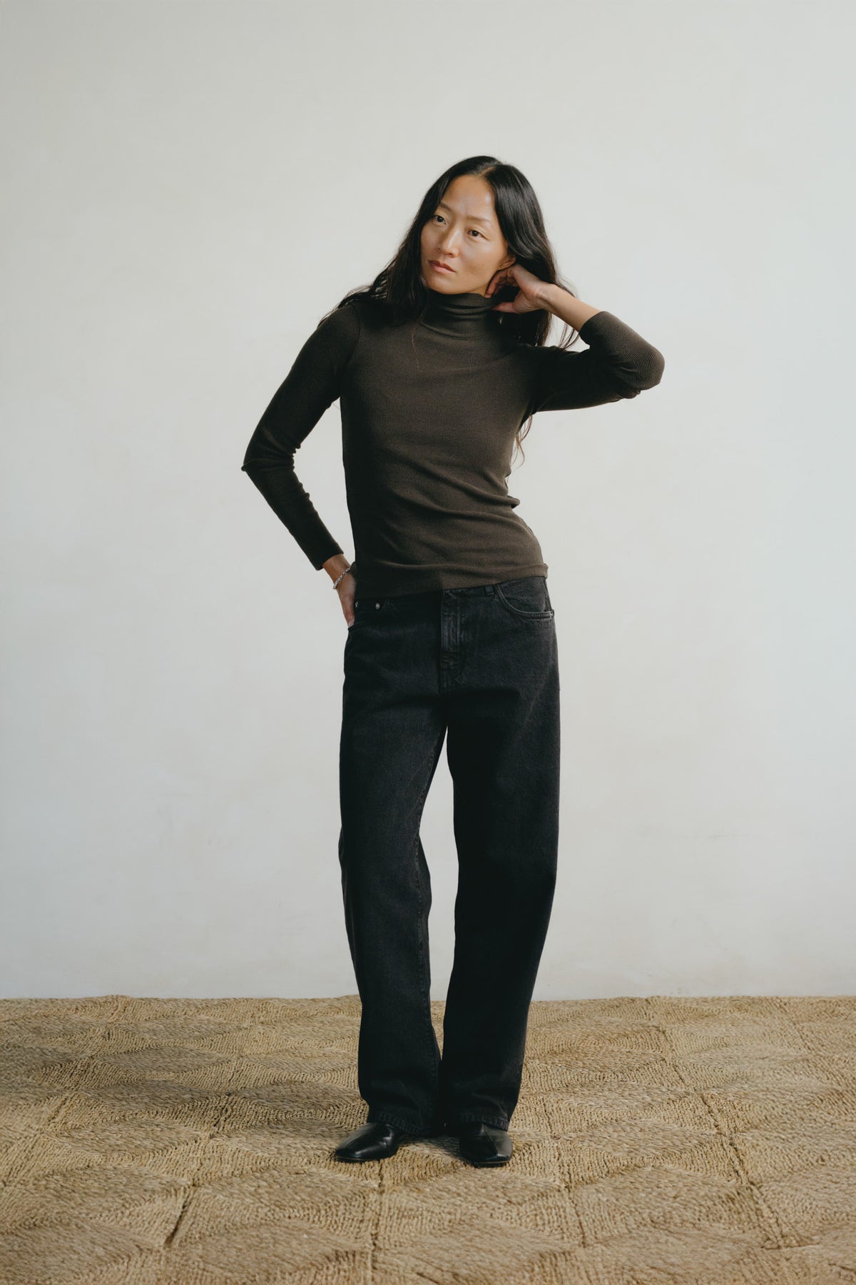 A woman modeling a dark brown ribbed turtleneck sweater and black wide-leg jeans against a minimalist off-white wall and textured beige floor.