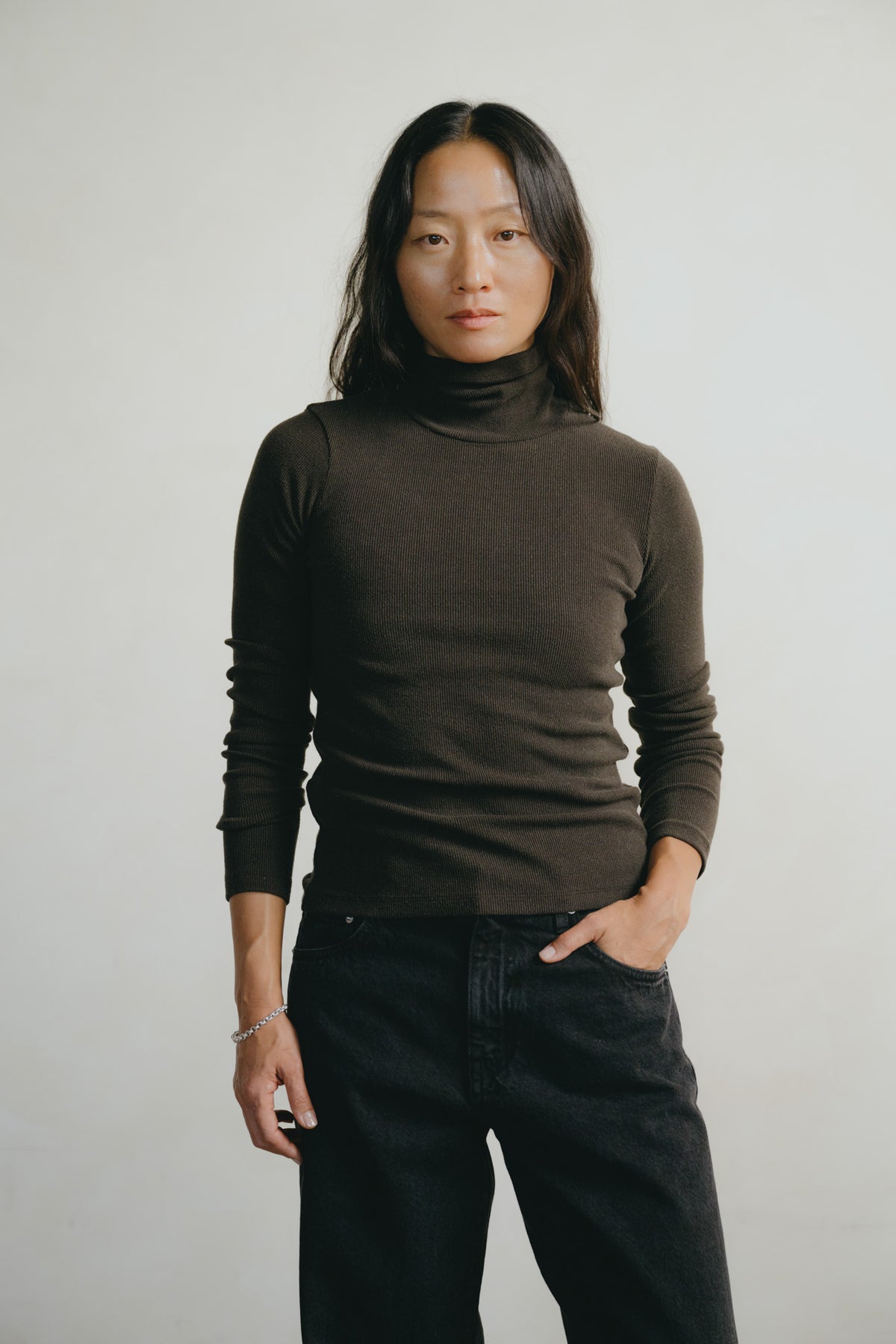 A woman modeling a dark brown ribbed turtleneck sweater and black jeans against a plain light gray studio background.