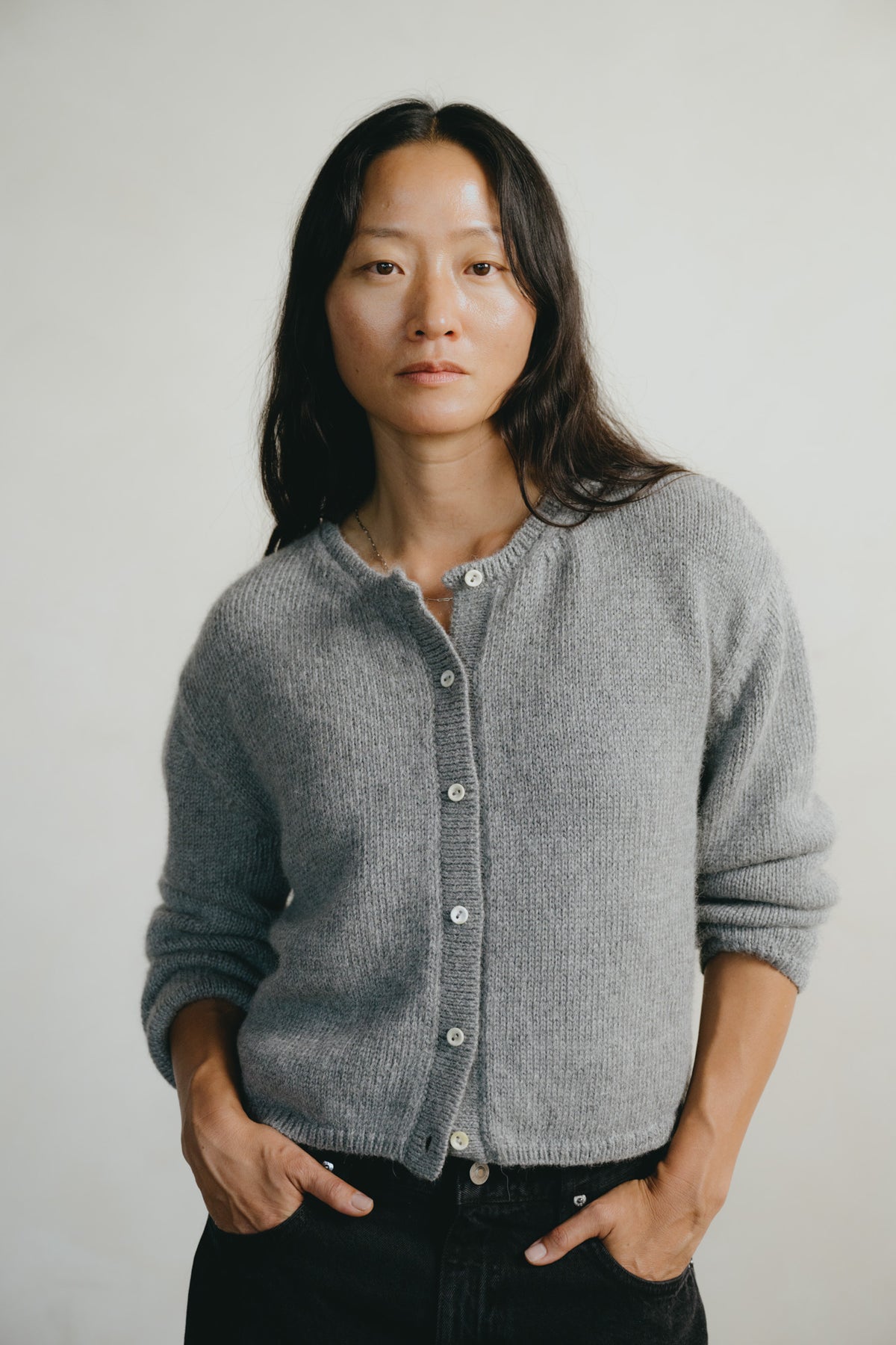 A woman with long dark hair wearing a gray ribbed knit cardigan with silver buttons, paired with black pants, standing against a plain light gray background.