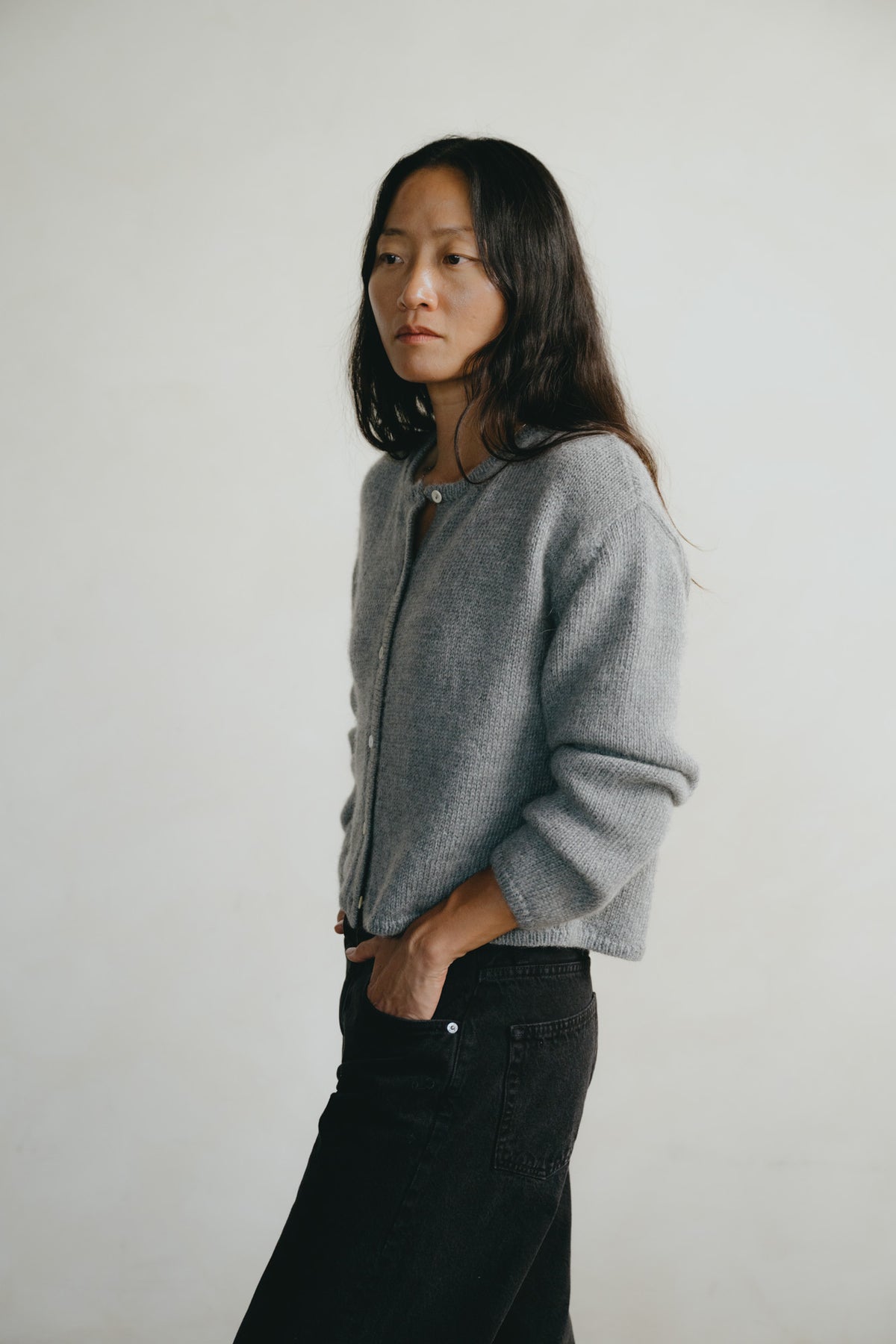A woman with long dark hair stands in profile wearing a light gray knit zip-up cardigan and black jeans against a plain light gray studio background.