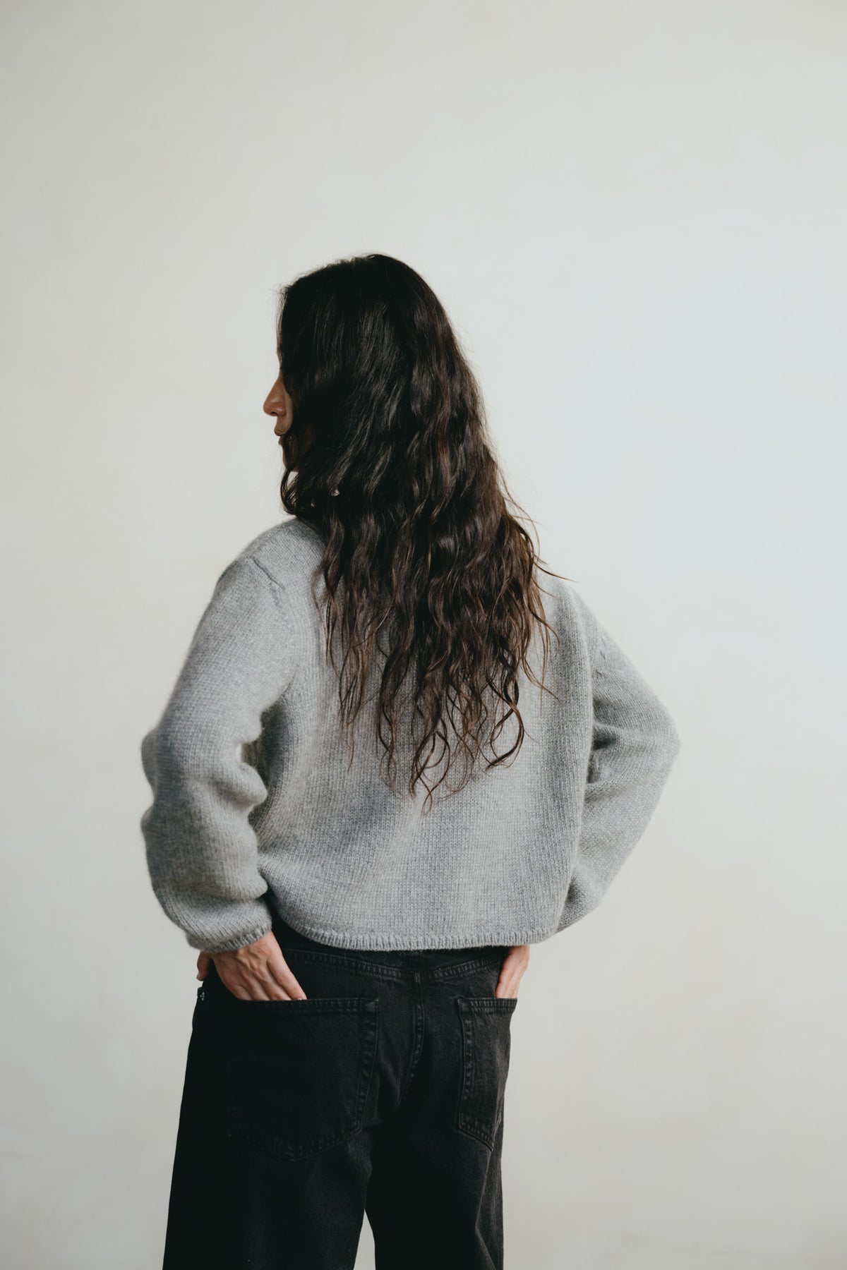 A woman with long, wavy dark hair is shown from behind, wearing a light gray fuzzy knit sweater and black jeans against a plain off-white background.
