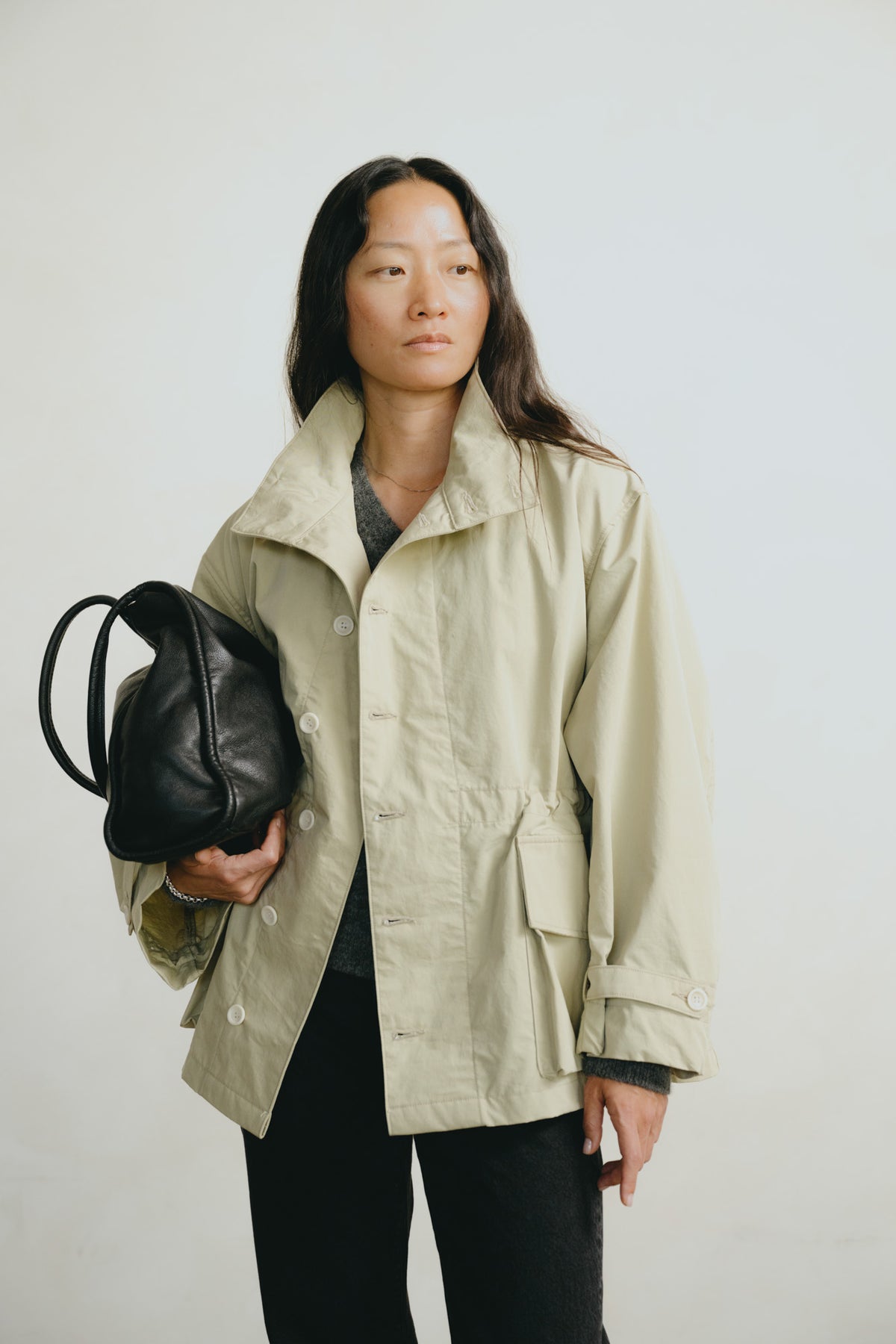 A woman modeling a light beige utility jacket, holding a black leather shoulder bag, against a plain light gray studio background.
