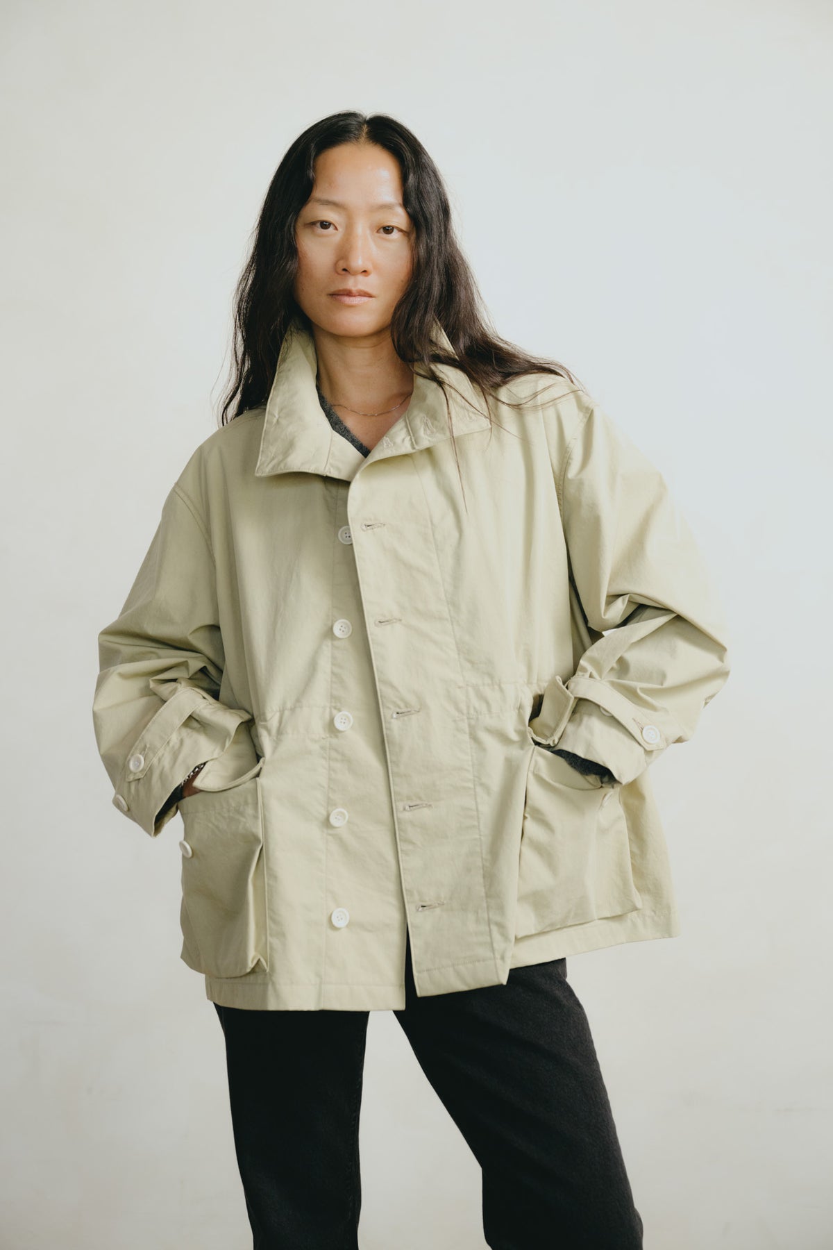 A model wearing an oversized light beige utility jacket with front buttons and large patch pockets, paired with black pants, against a plain light gray background.