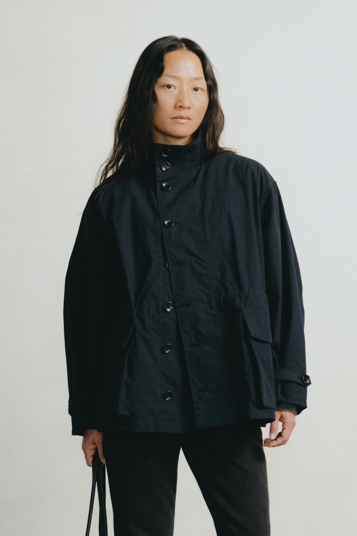 A model wearing a black high-collar utility jacket with front buttons and side pockets, paired with black pants, standing against a light gray studio background.