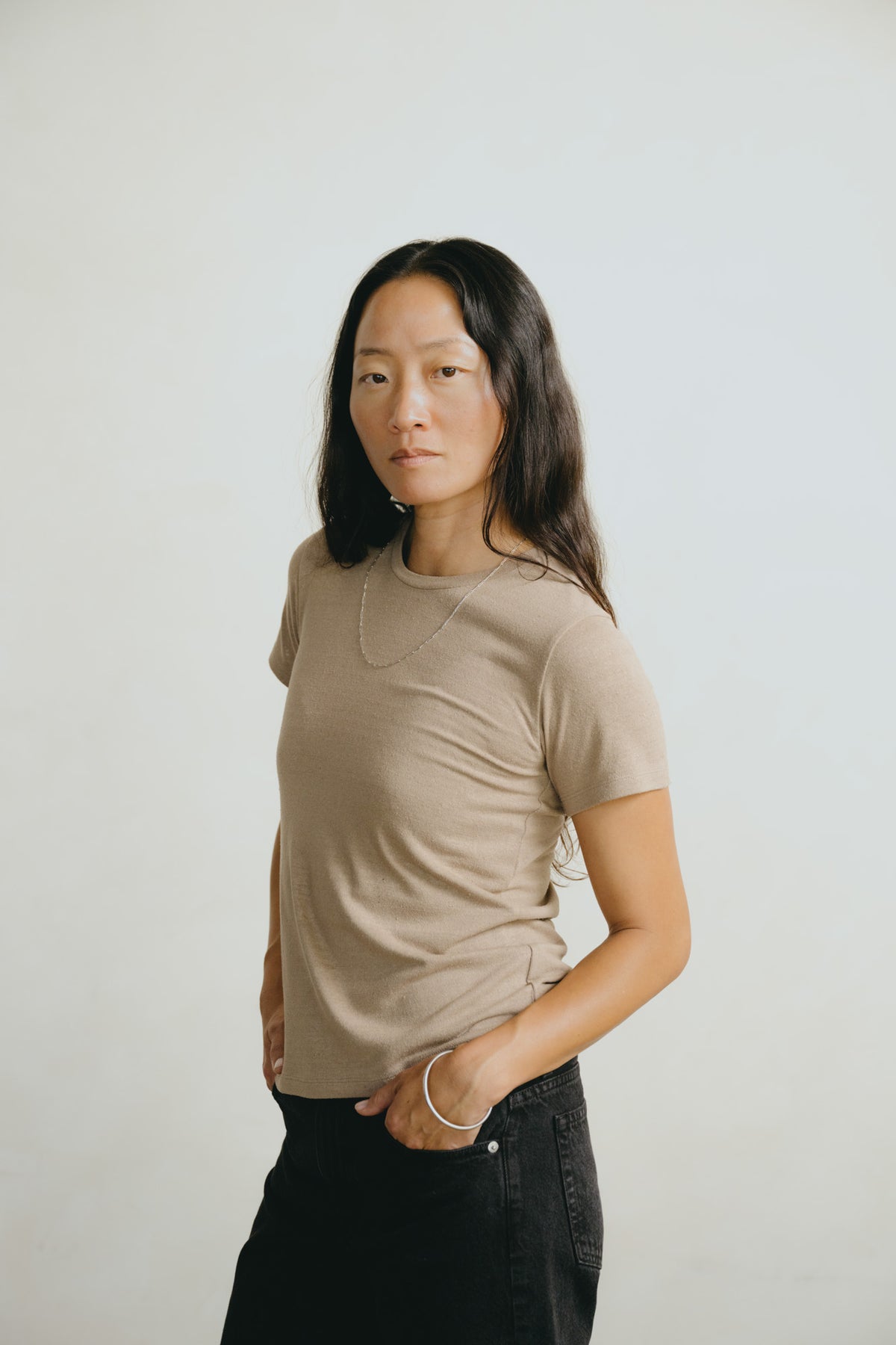 A woman with long dark hair wearing a beige short-sleeve crewneck t-shirt and black jeans, standing against a plain light gray background.