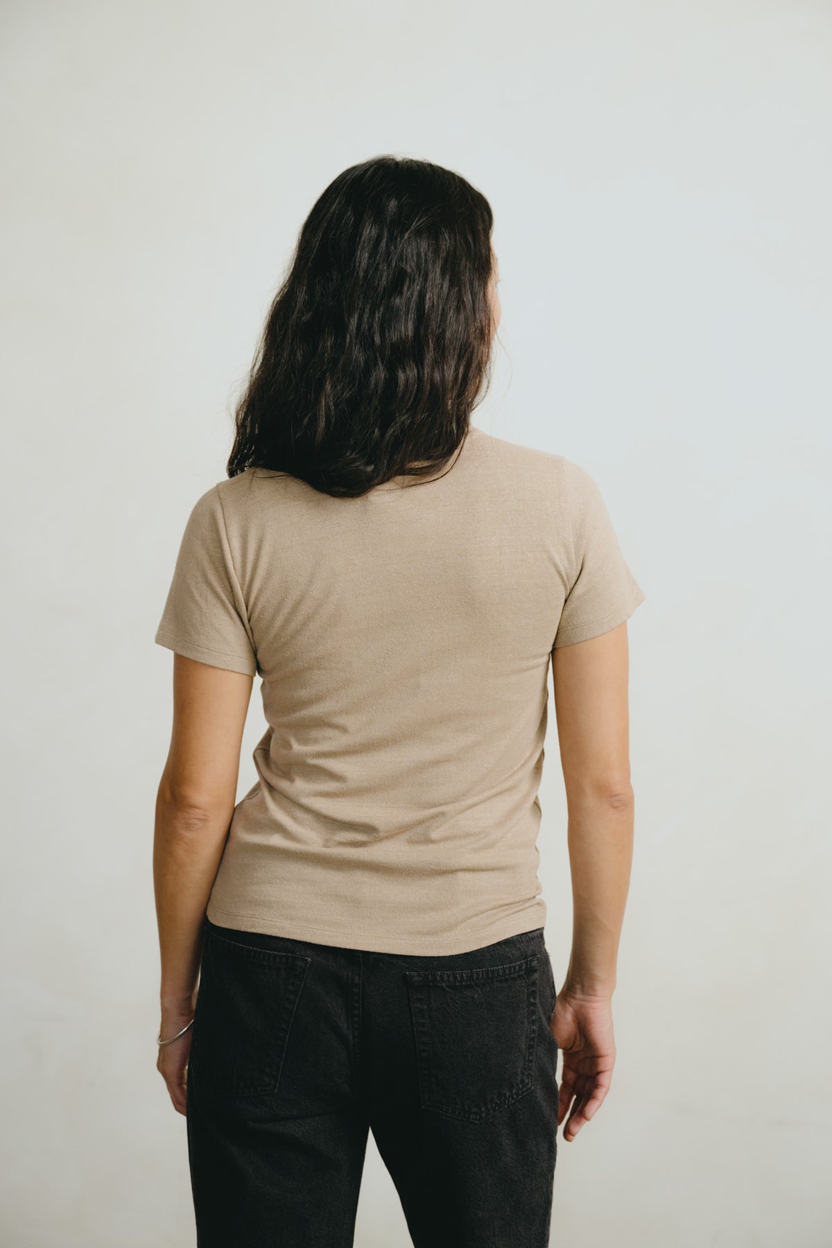 A woman with long dark hair is shown from behind wearing a light beige short-sleeve t-shirt and black jeans against a plain light gray background.