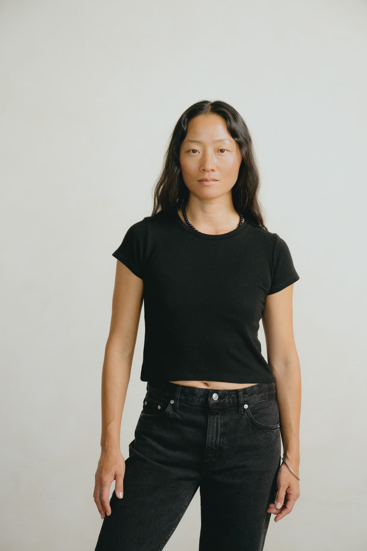 A woman modeling a black short-sleeve cropped t-shirt and black jeans against a plain light gray studio background.