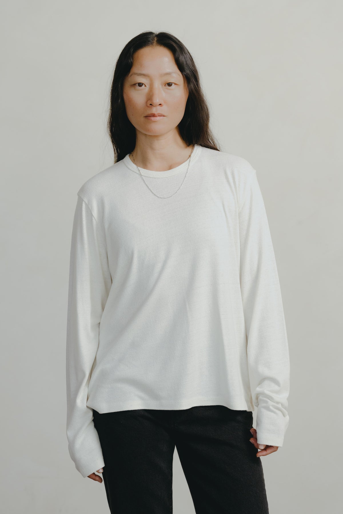 A woman wearing a white long-sleeve crewneck shirt and black pants, standing against a light gray studio background.