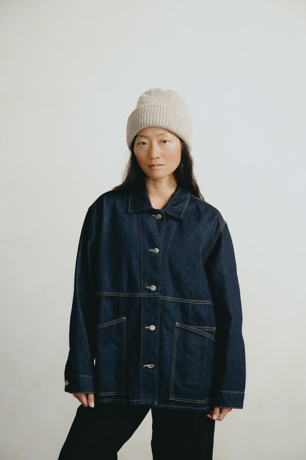 A model wearing a dark indigo denim jacket with contrast light-colored stitching, paired with black pants and a beige knit beanie, standing against a plain off-white background.