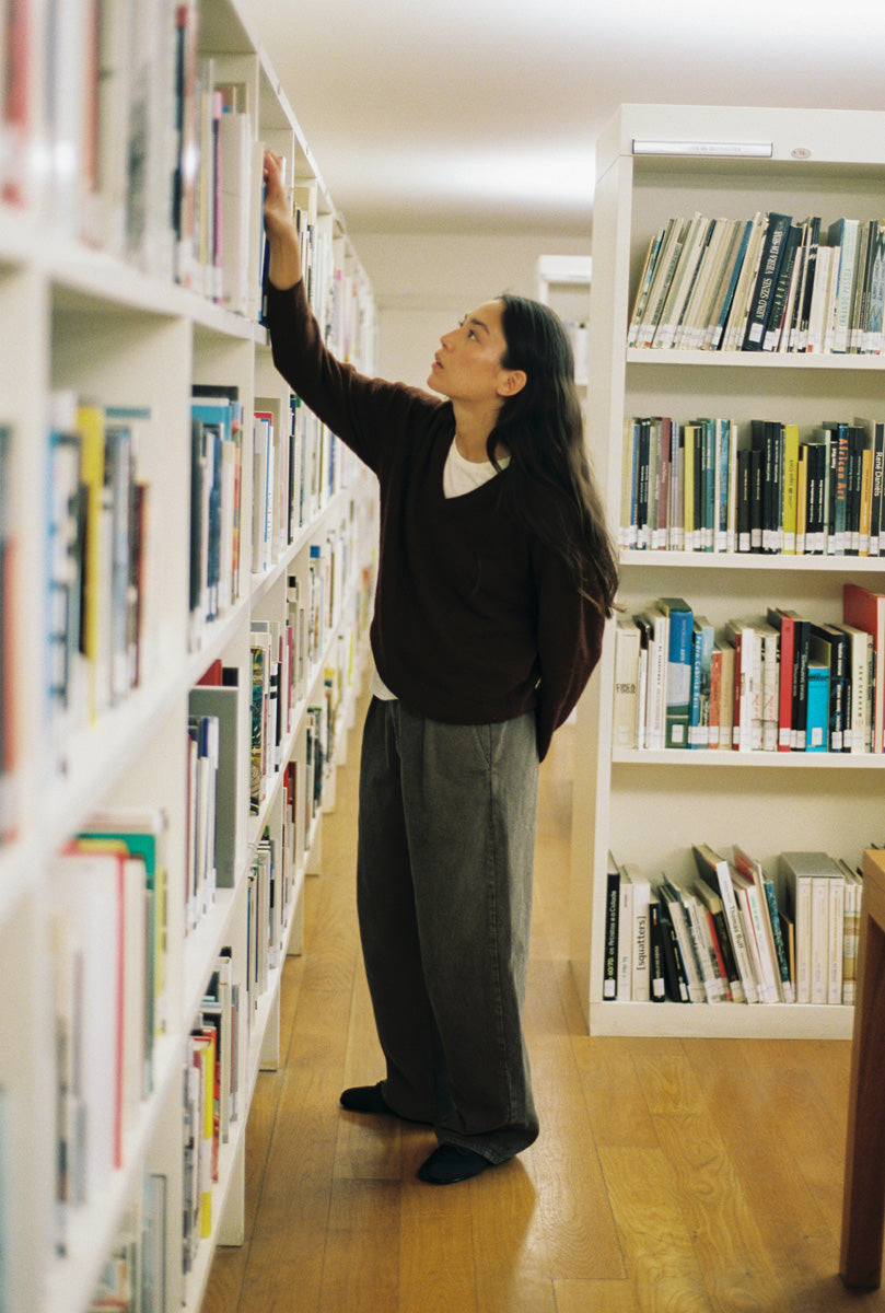 A woman with long dark hair stands in a library aisle between white bookshelves, reaching up to pick a book. She is wearing a dark brown sweater, loose gray trousers, and black socks on a light wood floor.