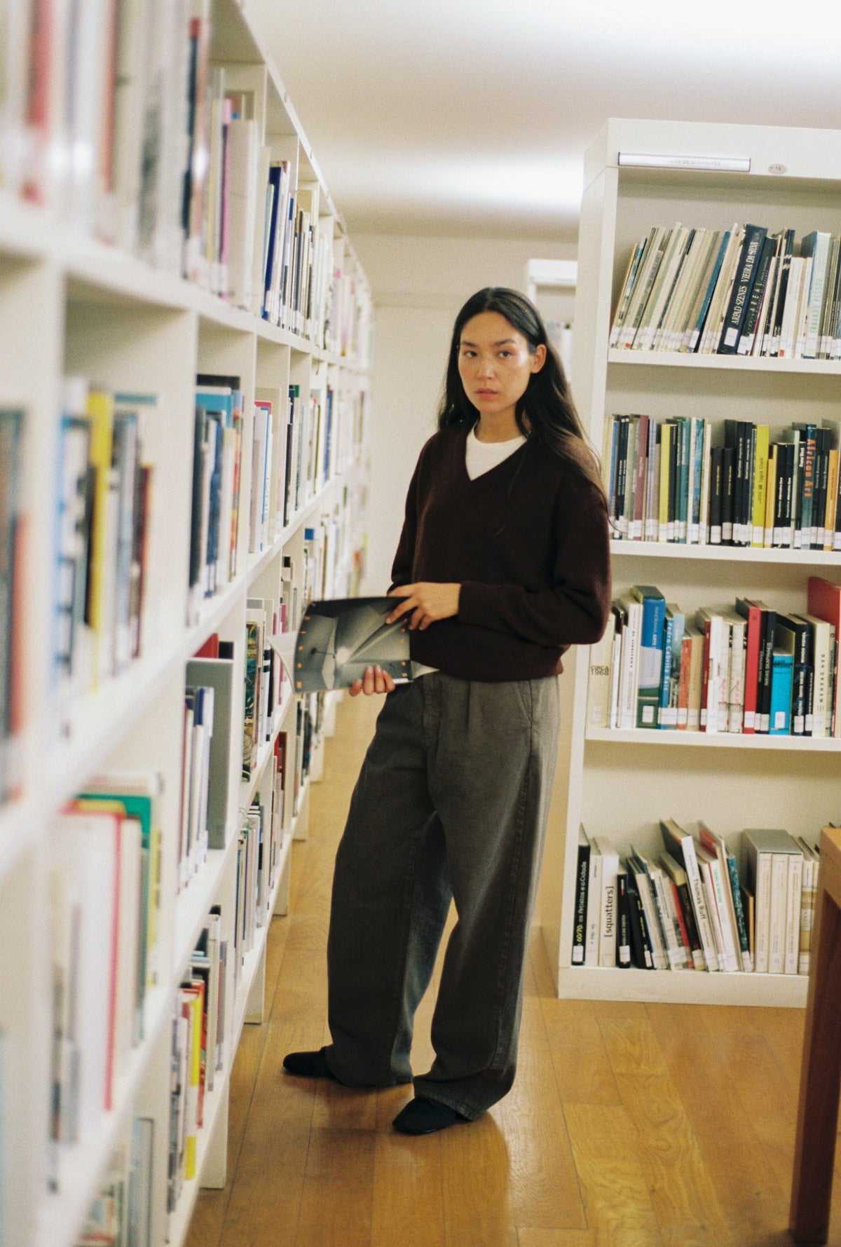 A woman stands in a library aisle between white bookshelves, holding a tablet. She is wearing a dark brown v-neck sweater over a white shirt, loose dark gray trousers, and black socks.