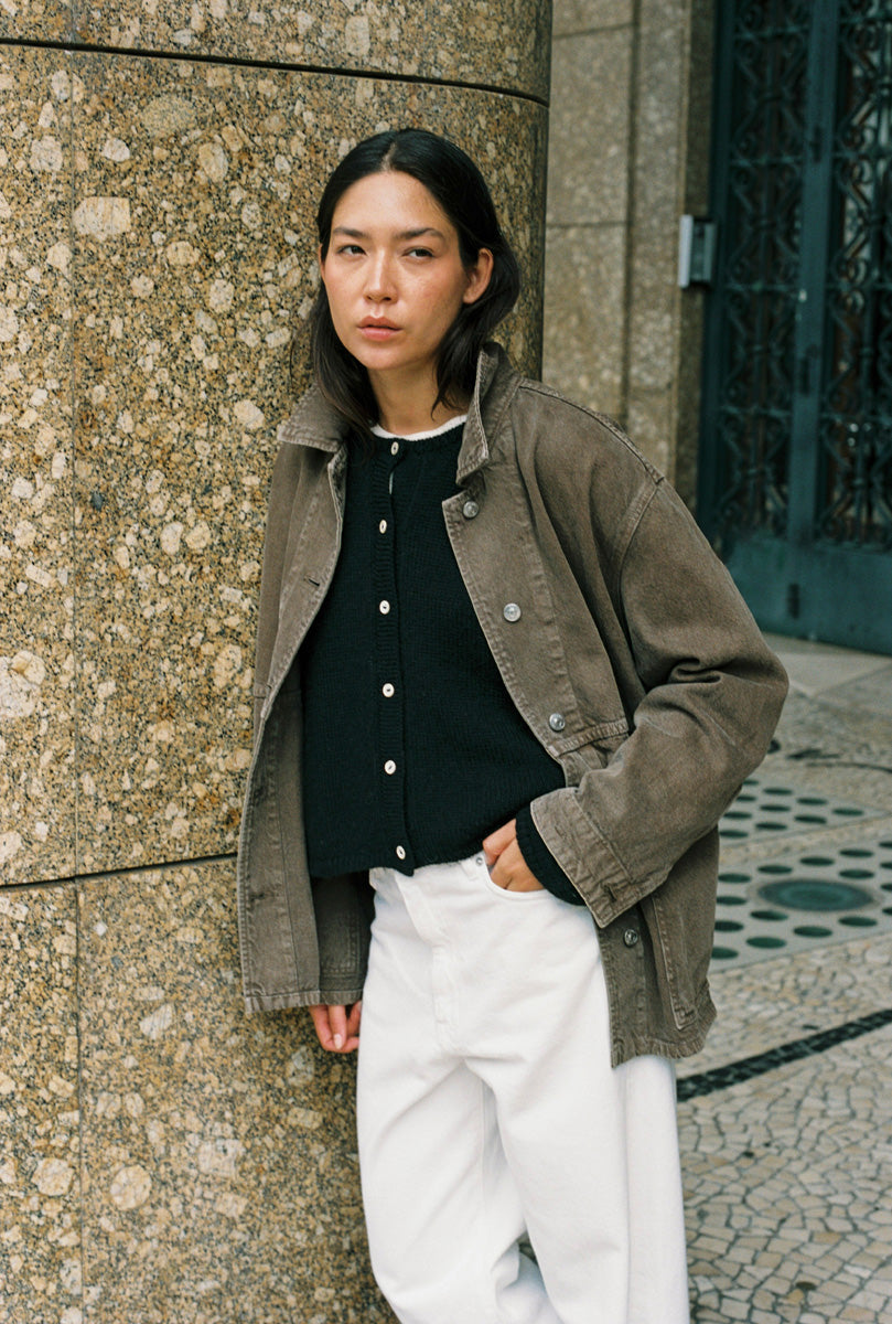 A woman stands outdoors against a textured stone wall, wearing an oversized brown jacket over a black button-front top and white pants.